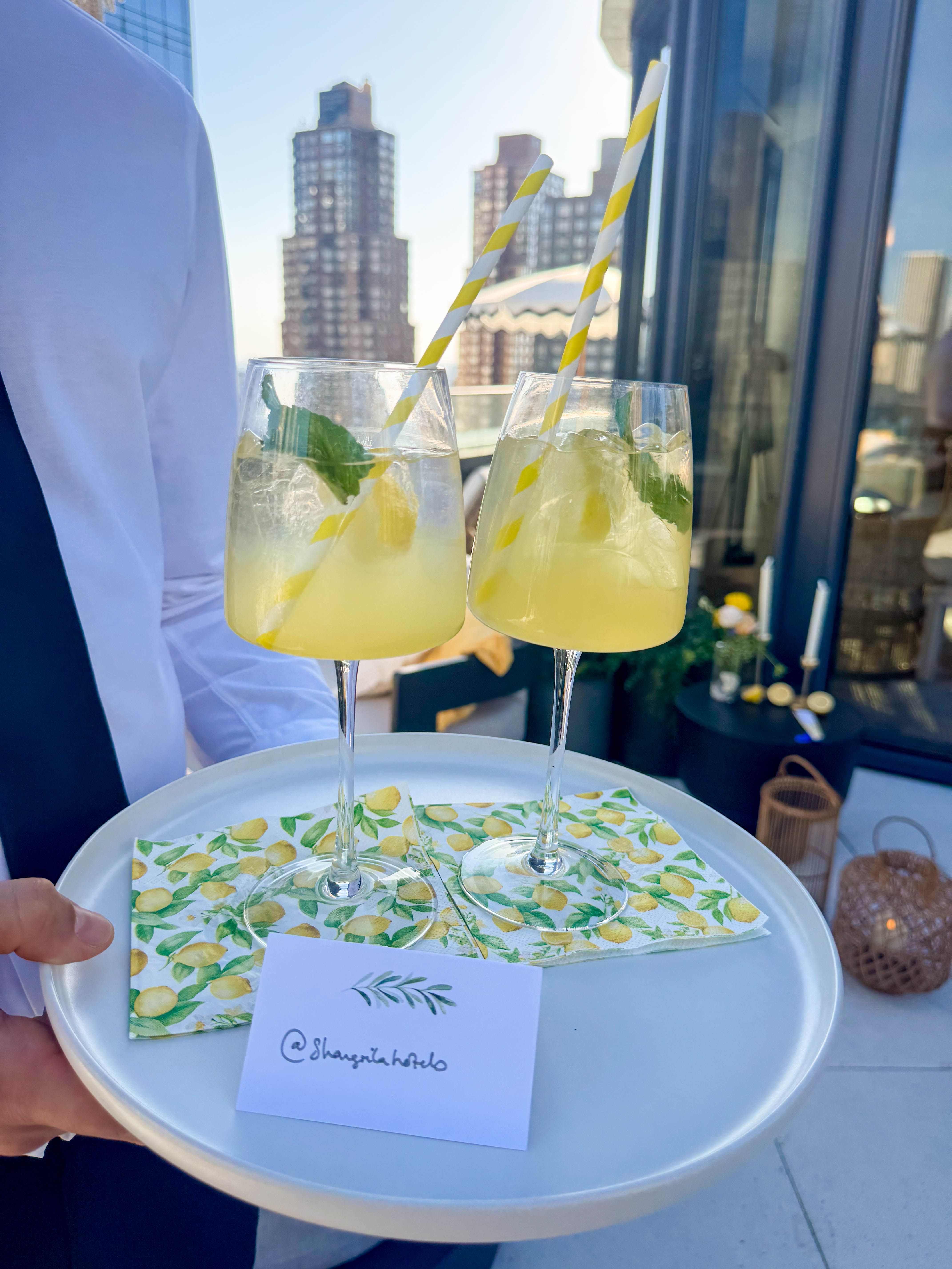 A person holds a tray with two lemon cocktails, garnished with mint and striped straws, on lemon-patterned napkins, next to a card for "@shangrila_hotels," with a city skyline in the background.