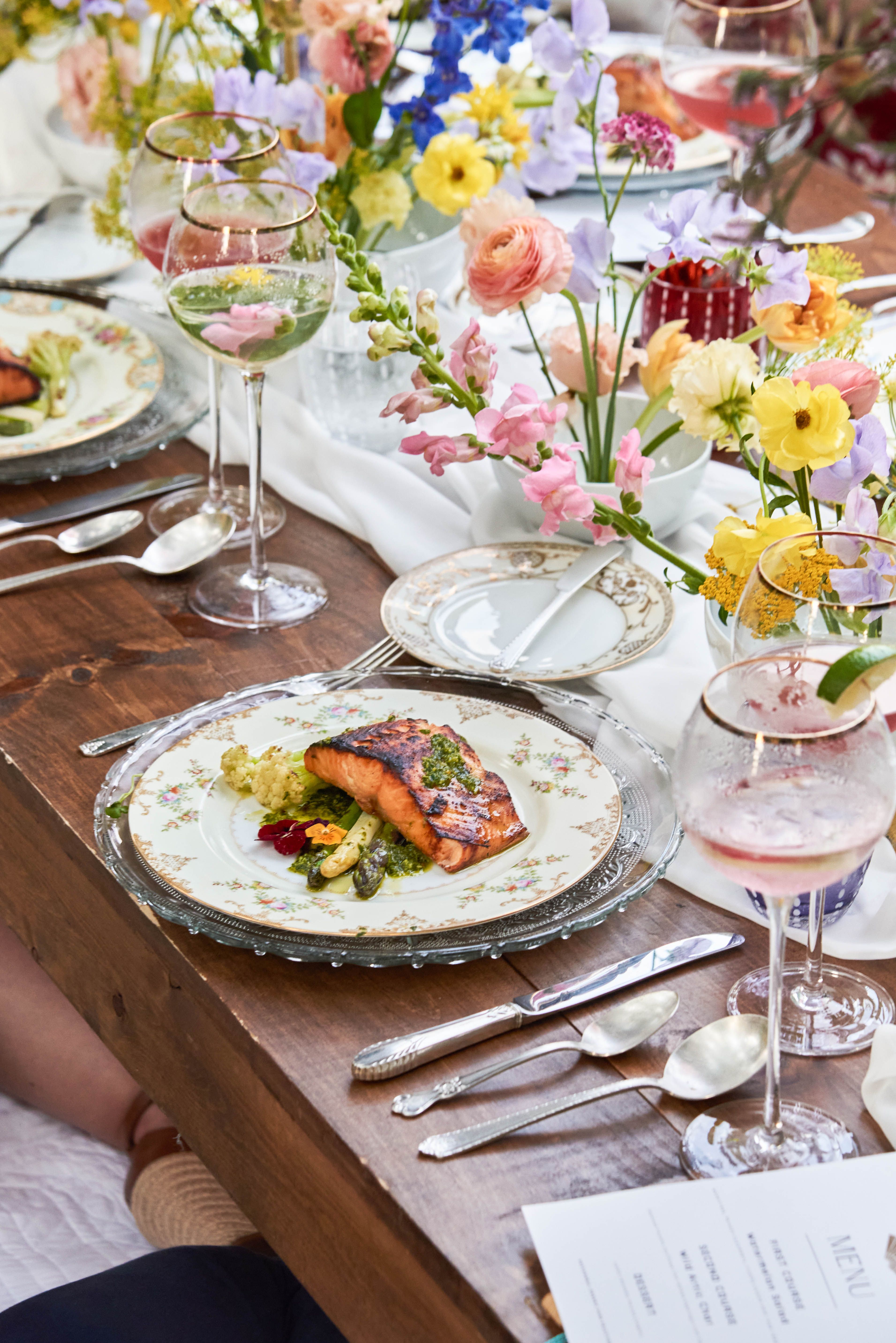 An elegant outdoor dining table made of wood, set with plates of grilled salmon and vegetables, vibrant floral arrangements, and wine glasses.
