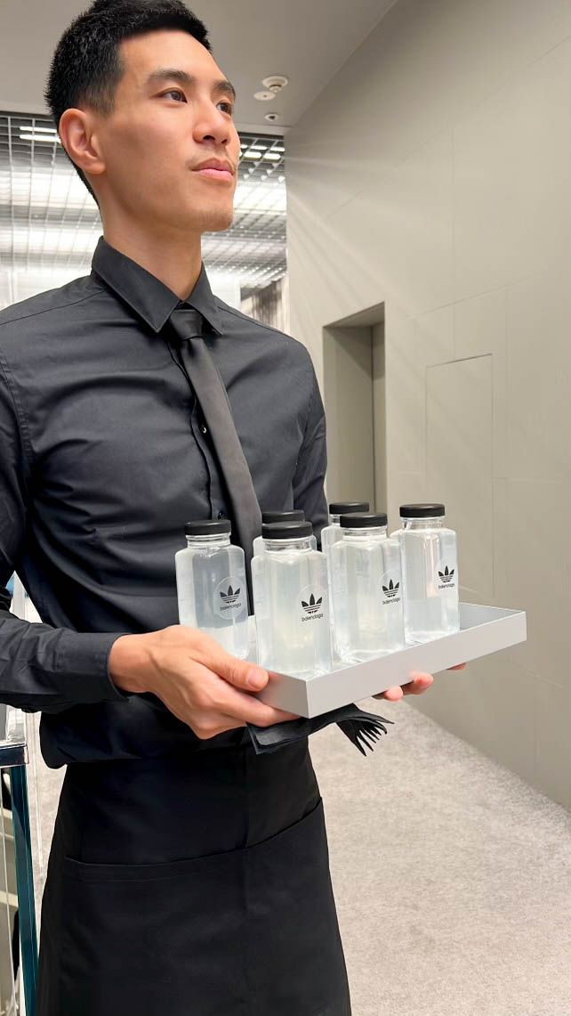 Server presenting branded bottled beverages on a tray during a luxury fashion brand experience event.