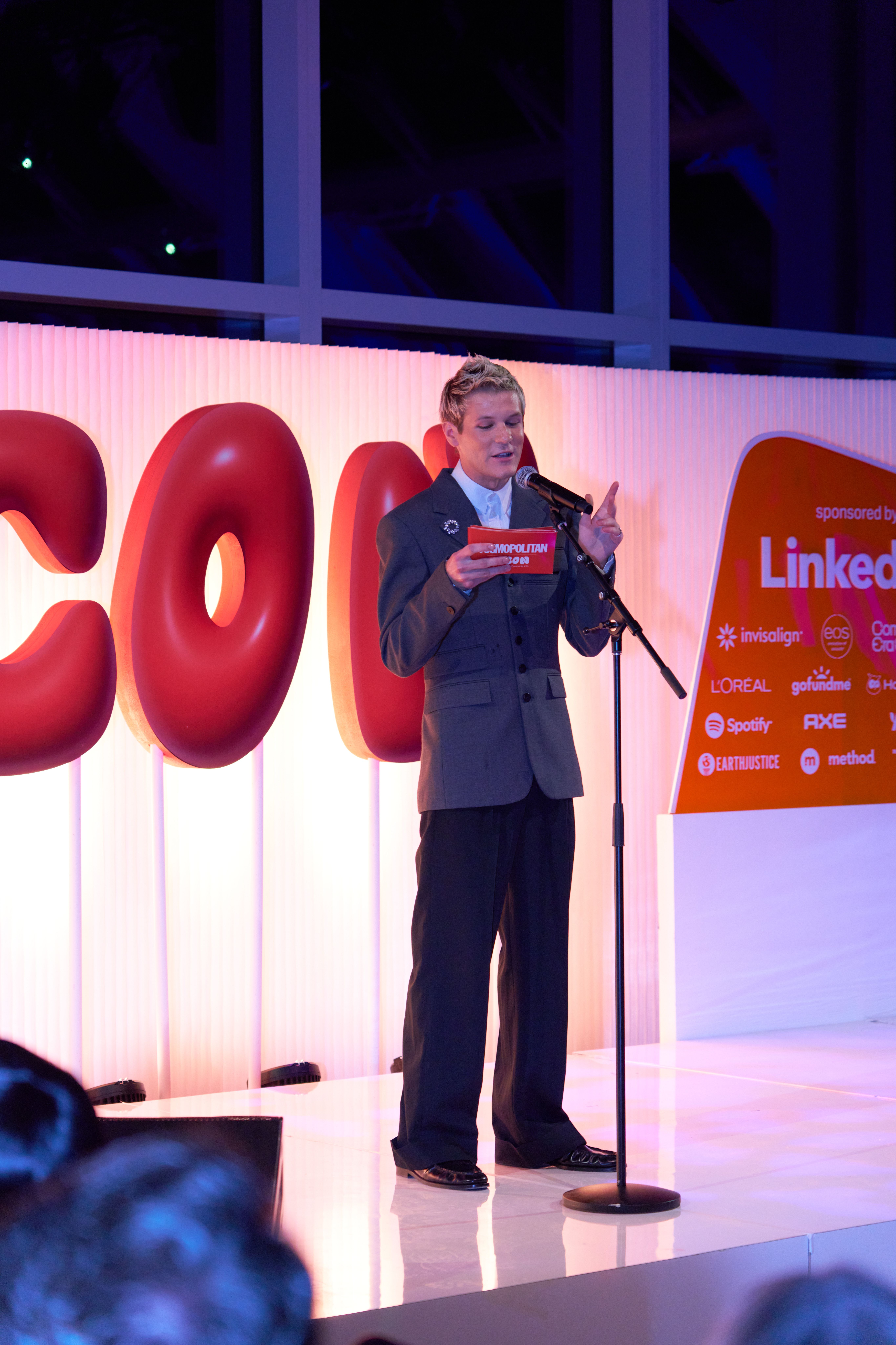 Speaker presenting on stage at a large corporate event reception with branded backdrop.