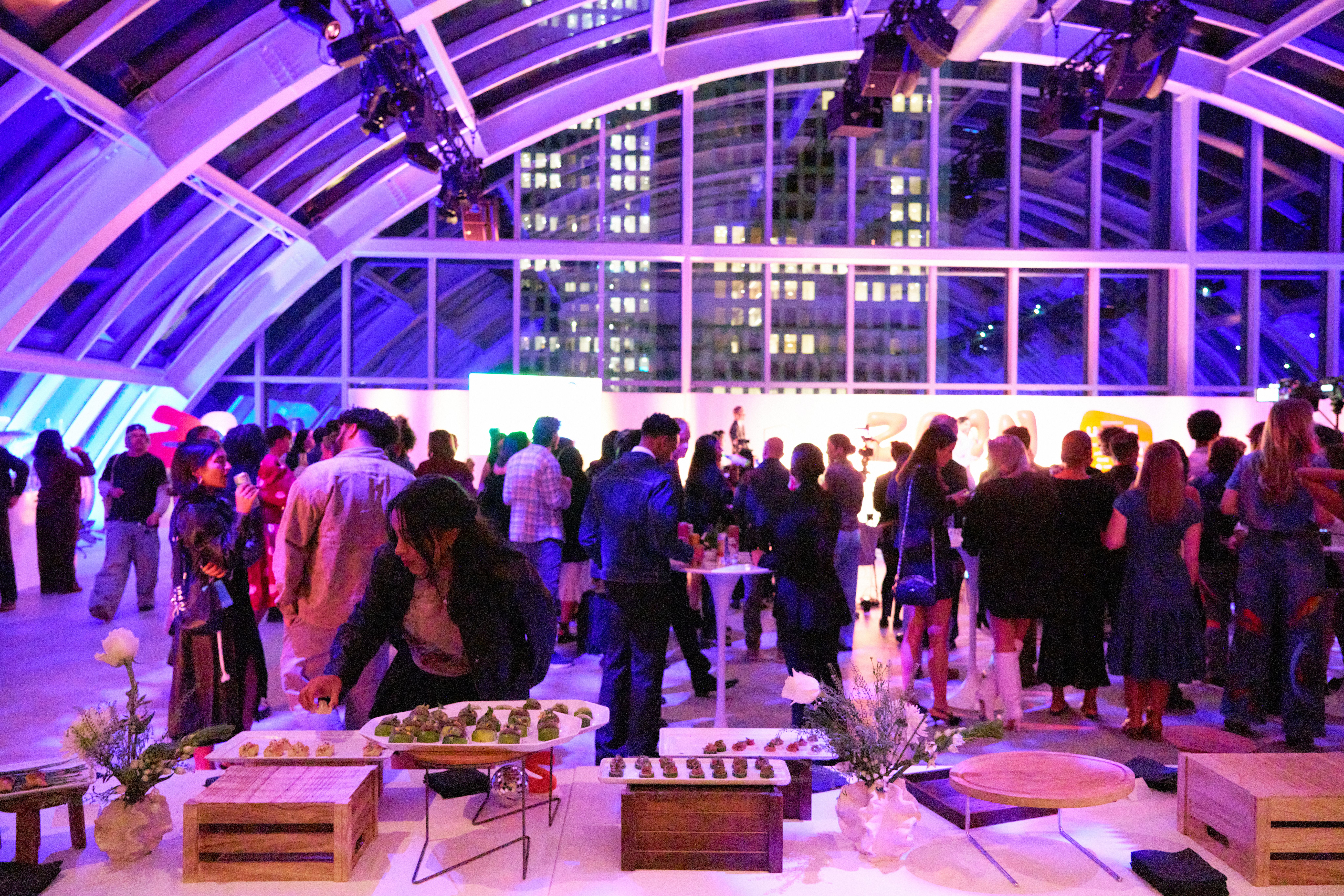 Many people mingle at a modern rooftop party with purple lighting, food displays, and city buildings visible through glass walls.