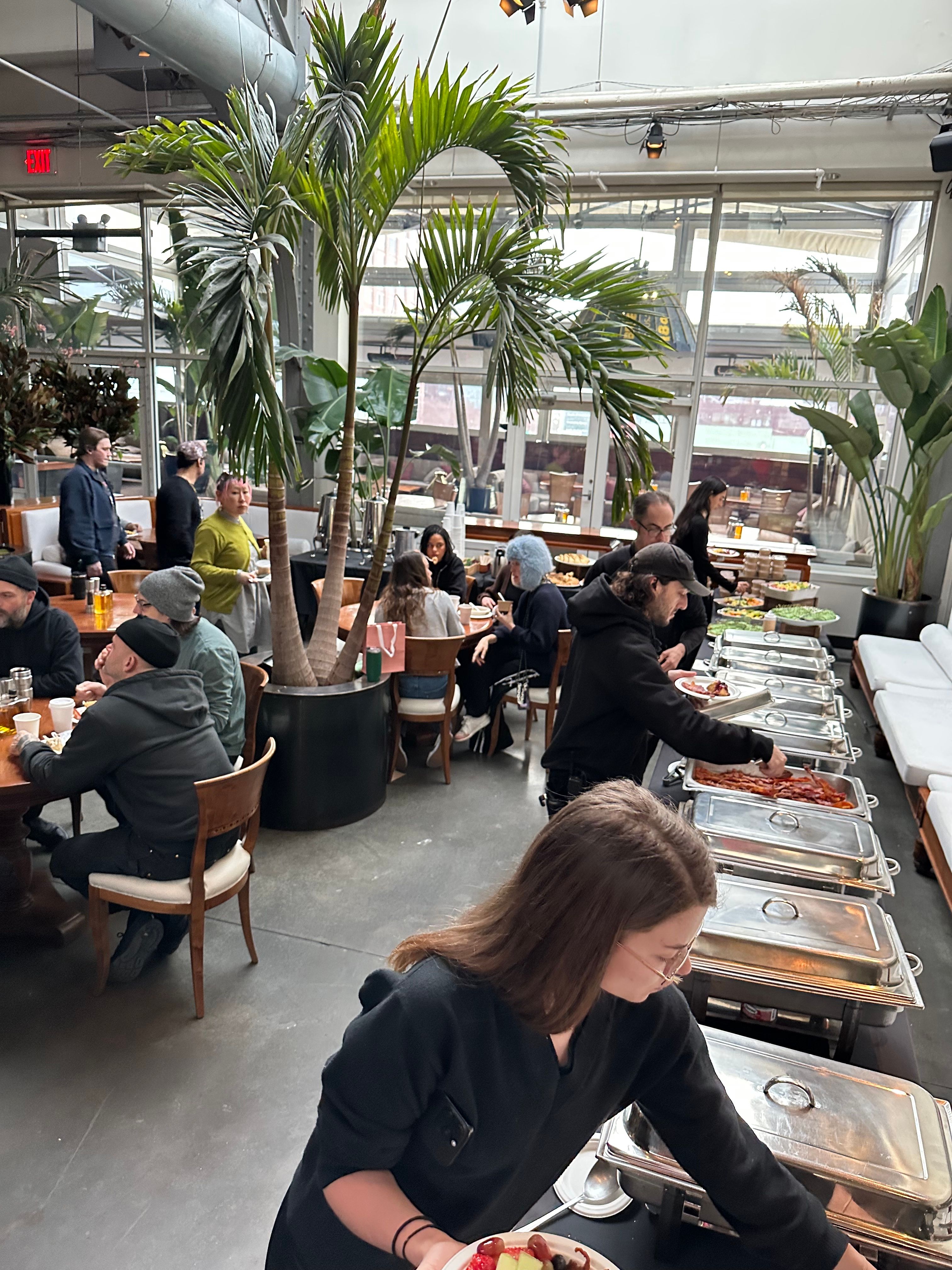 People serve themselves from a buffet line in a bright, plant-filled room with large windows.