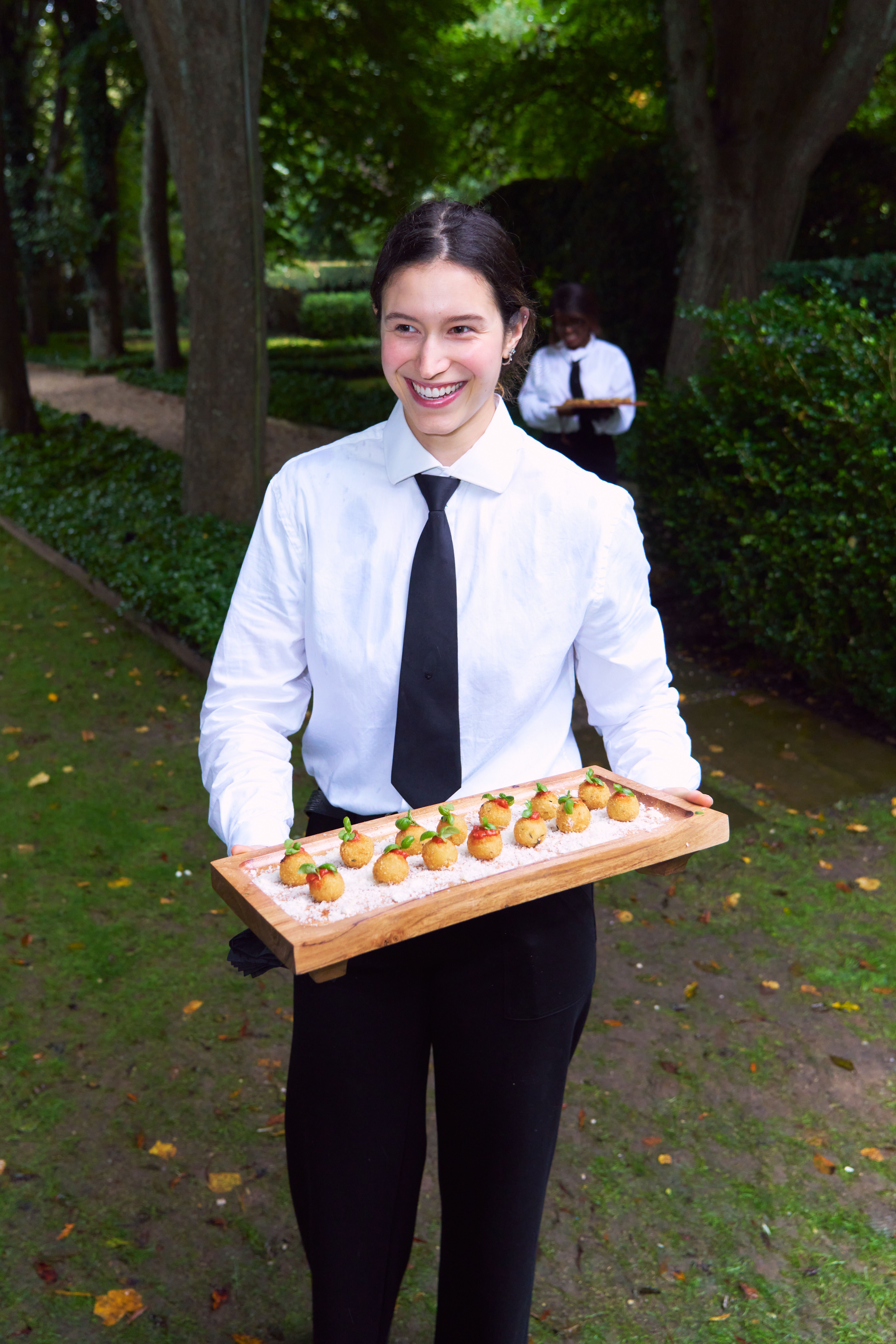 A smiling Hamptons estate wedding server in a white shirt and black tie holds a wooden tray of golden appetizers outdoors in a garden.