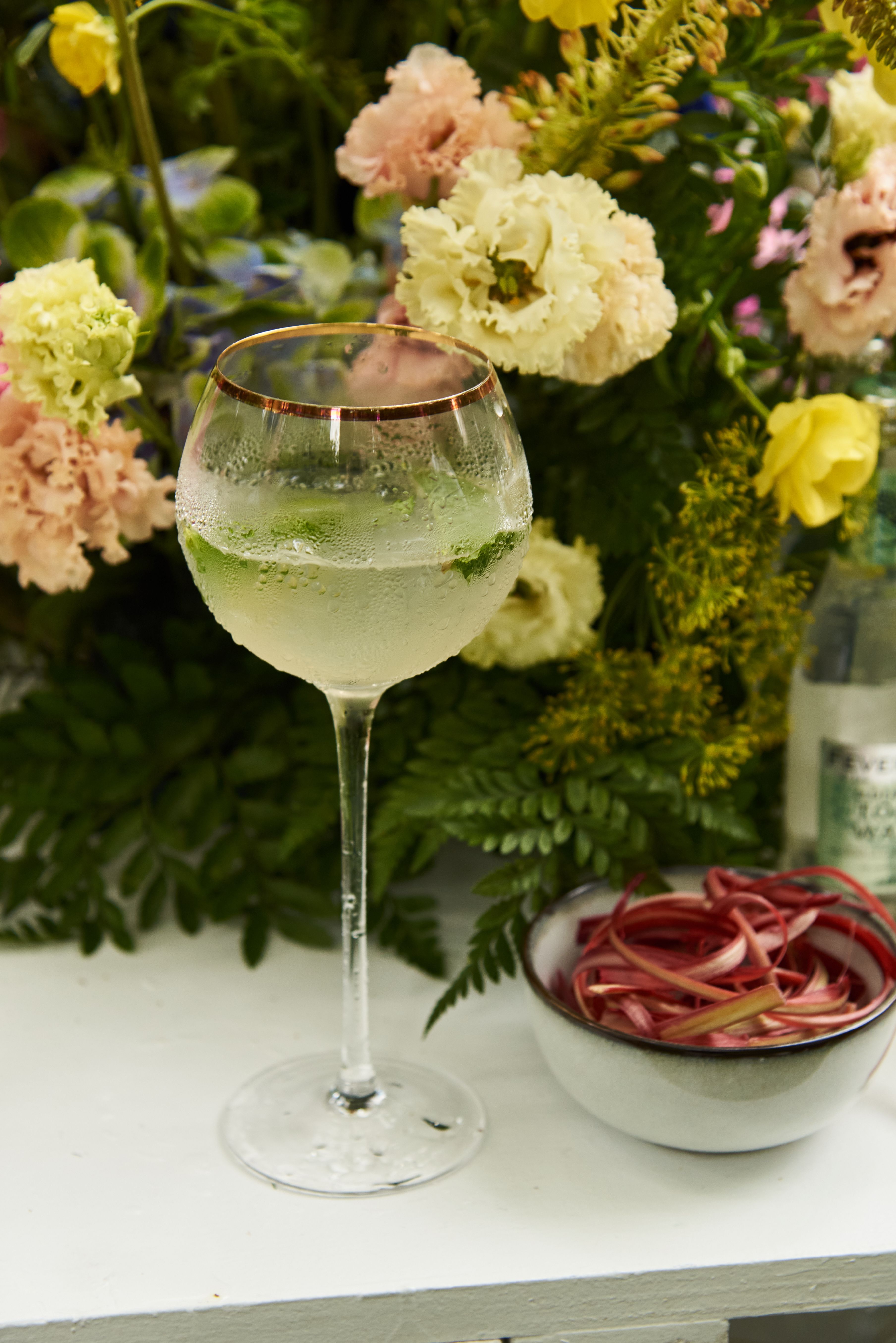 A frosty, gold-rimmed glass with a clear drink and mint leaves, next to a bowl of sliced rhubarb, against a vibrant floral background.