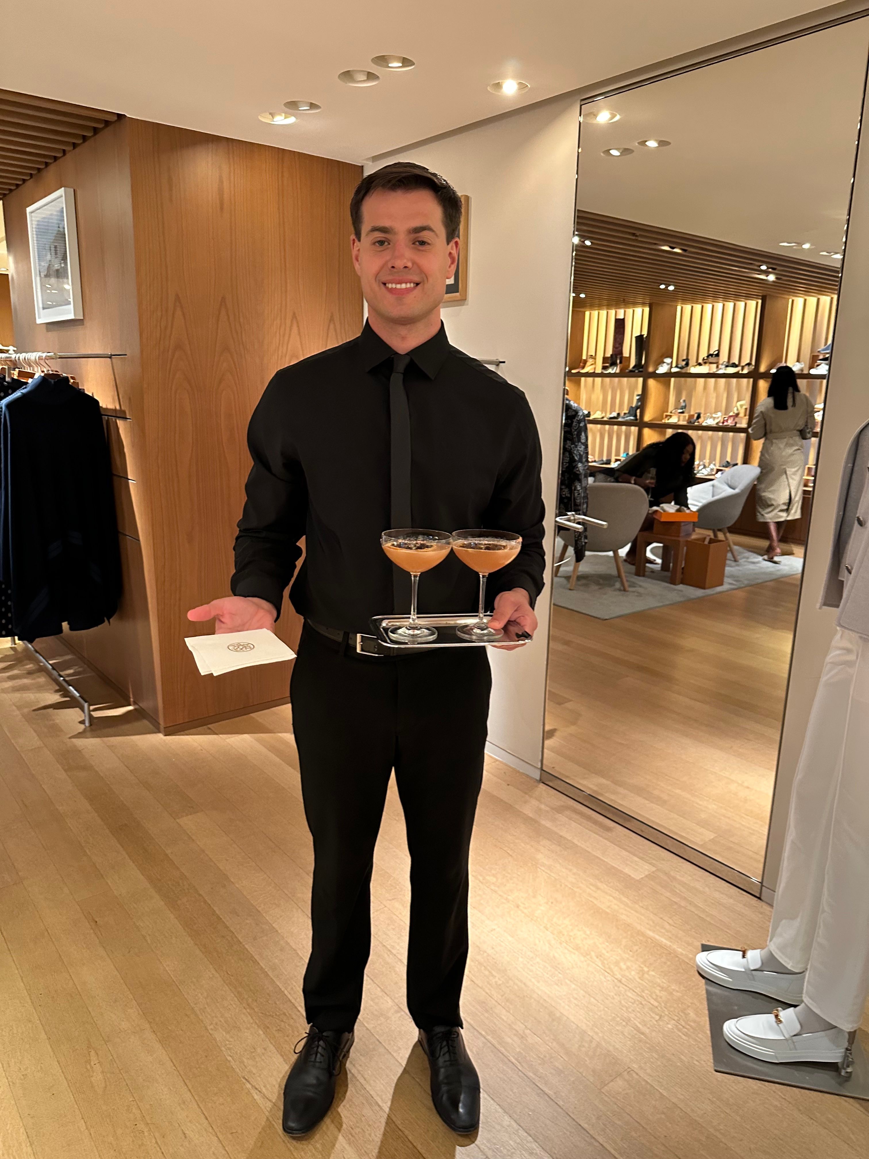 Hermes server in a black uniform holds a tray with two drinks and a white card in a stylish store.