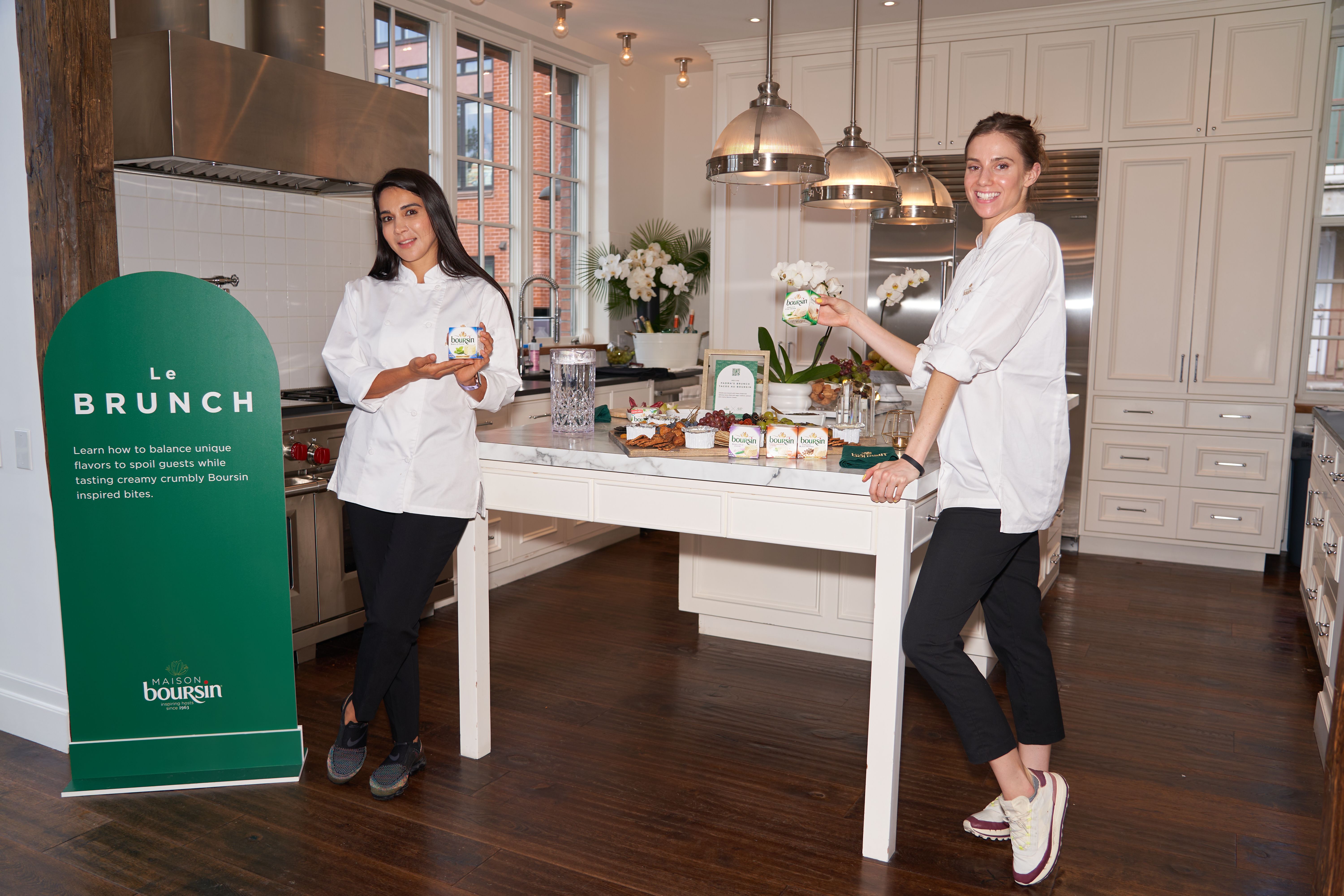 Two women in white tops display Boursin cheese in a bright kitchen with a "Le BRUNCH" sign and appetizers on a counter.