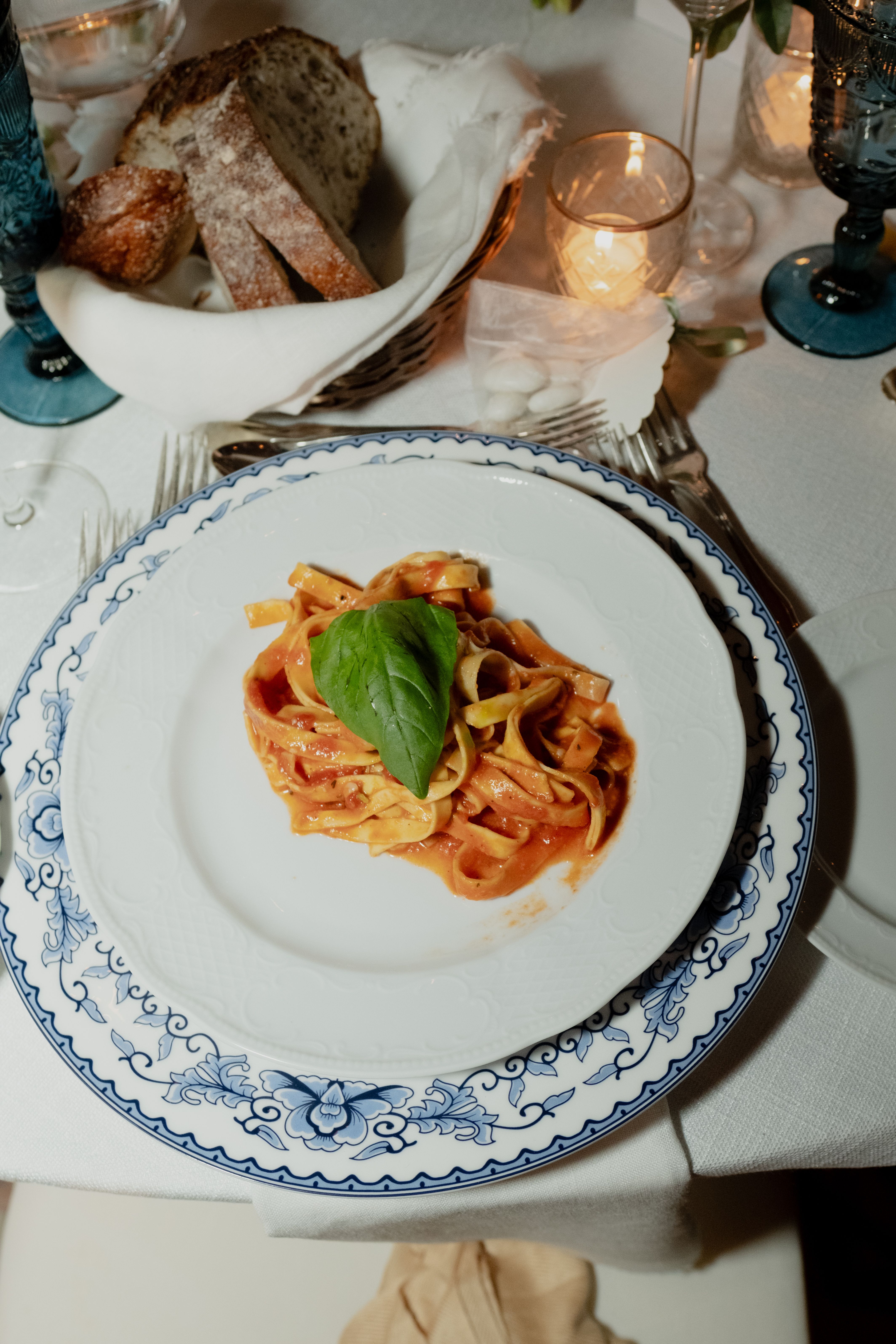 A plate of pasta with red sauce and a basil leaf sits on a dinner table with bread and a lit candle.