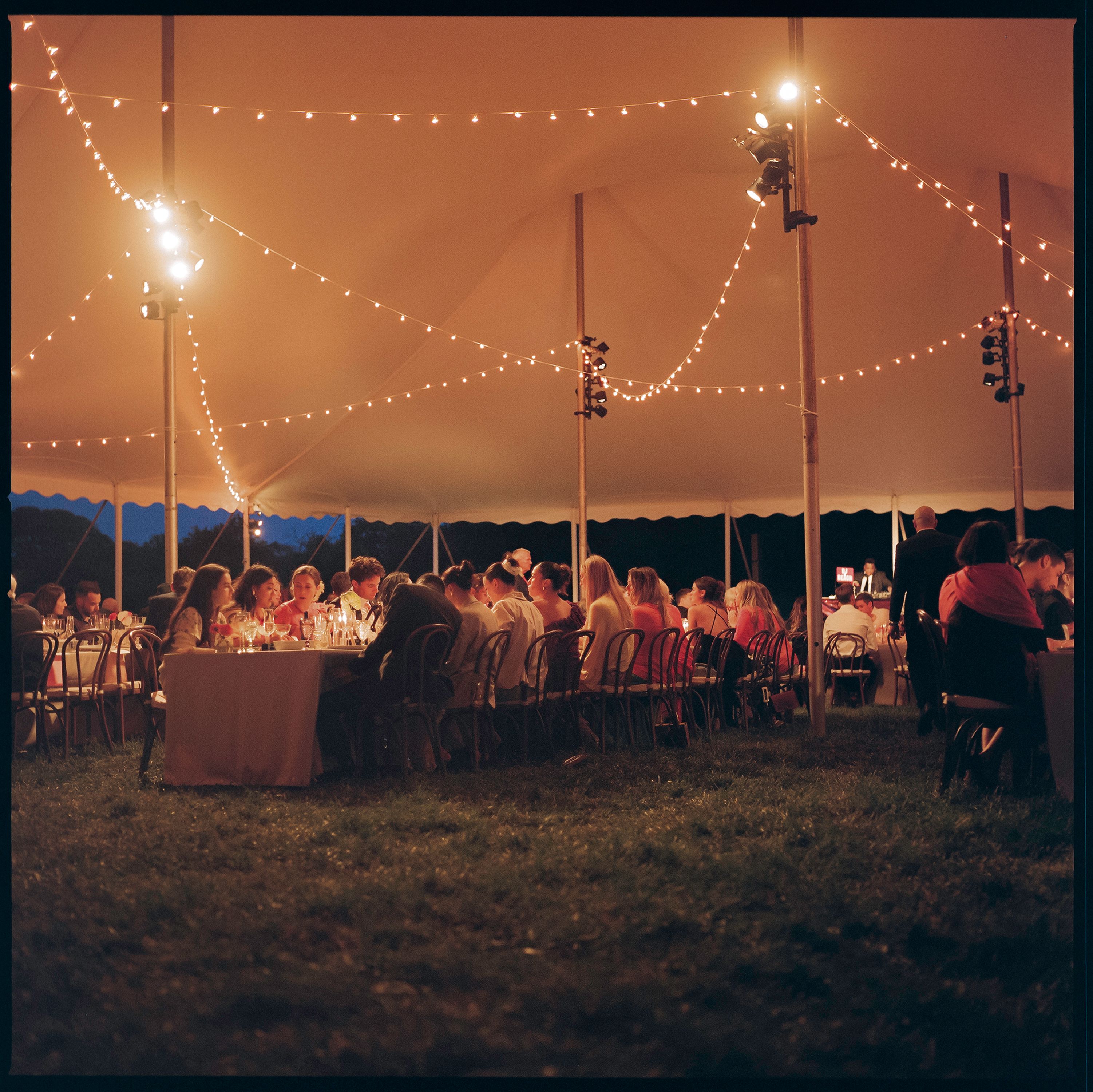 An outdoor evening event under a large white tent with string lights, showing guests seated at long tables on a grassy lawn.