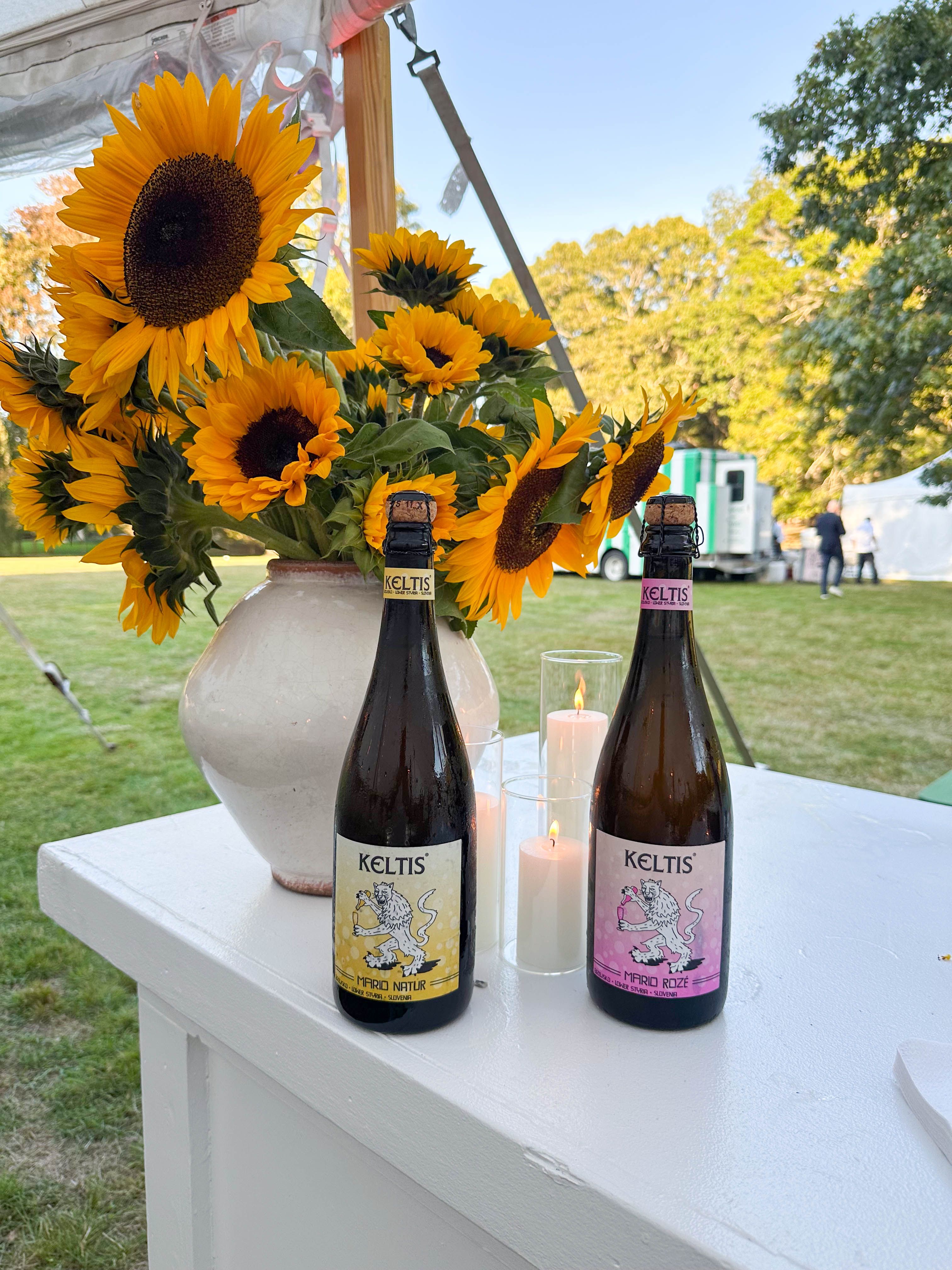 Two bottles of Keltis sparkling beverage, a vase of sunflowers, and lit candles on a white table at an outdoor wedding in the Hamptons..