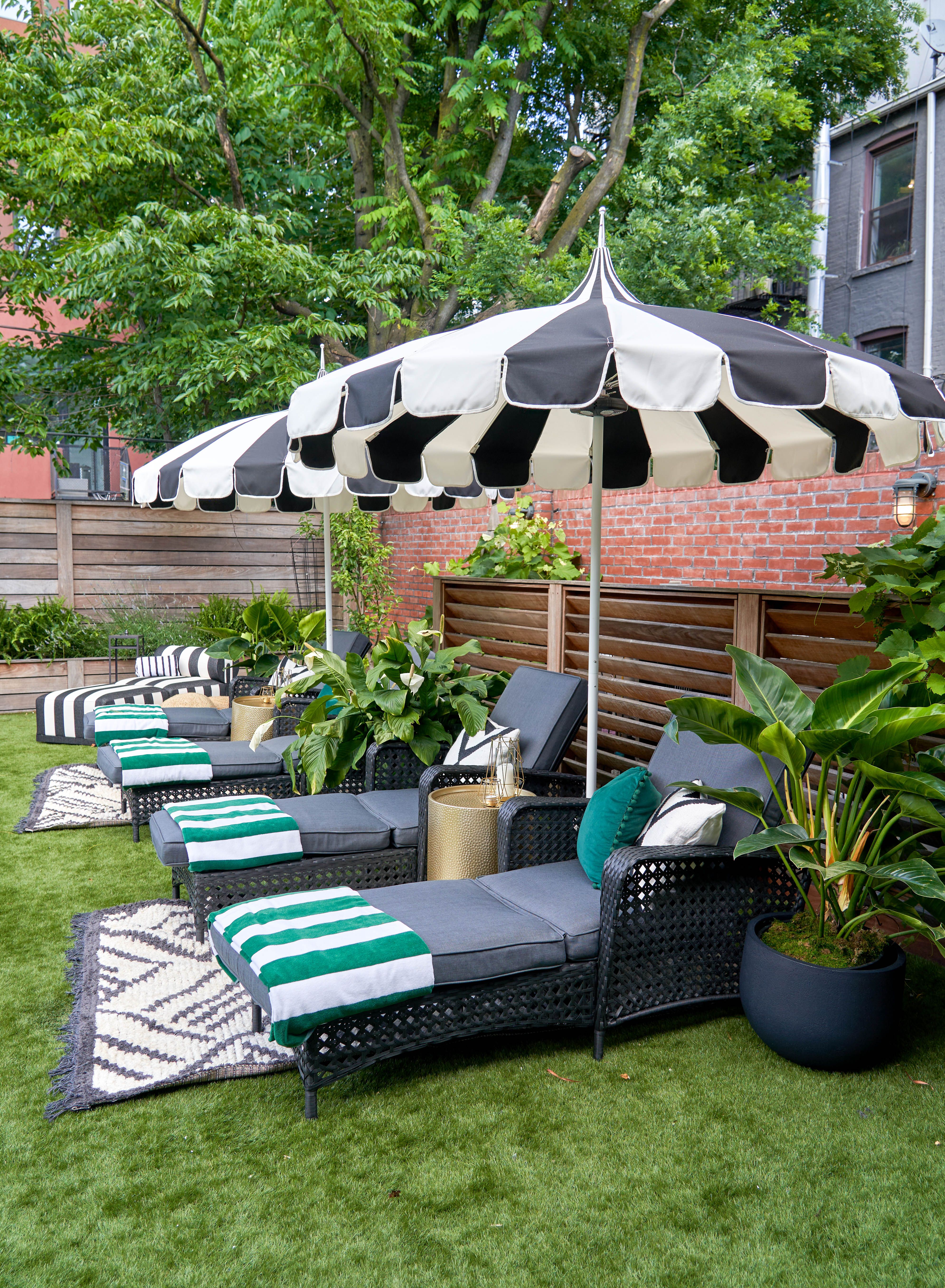 An outdoor lounge with black and white striped umbrellas shading dark gray chaise lounges with green and white blankets, set on artificial turf amidst lush plants.