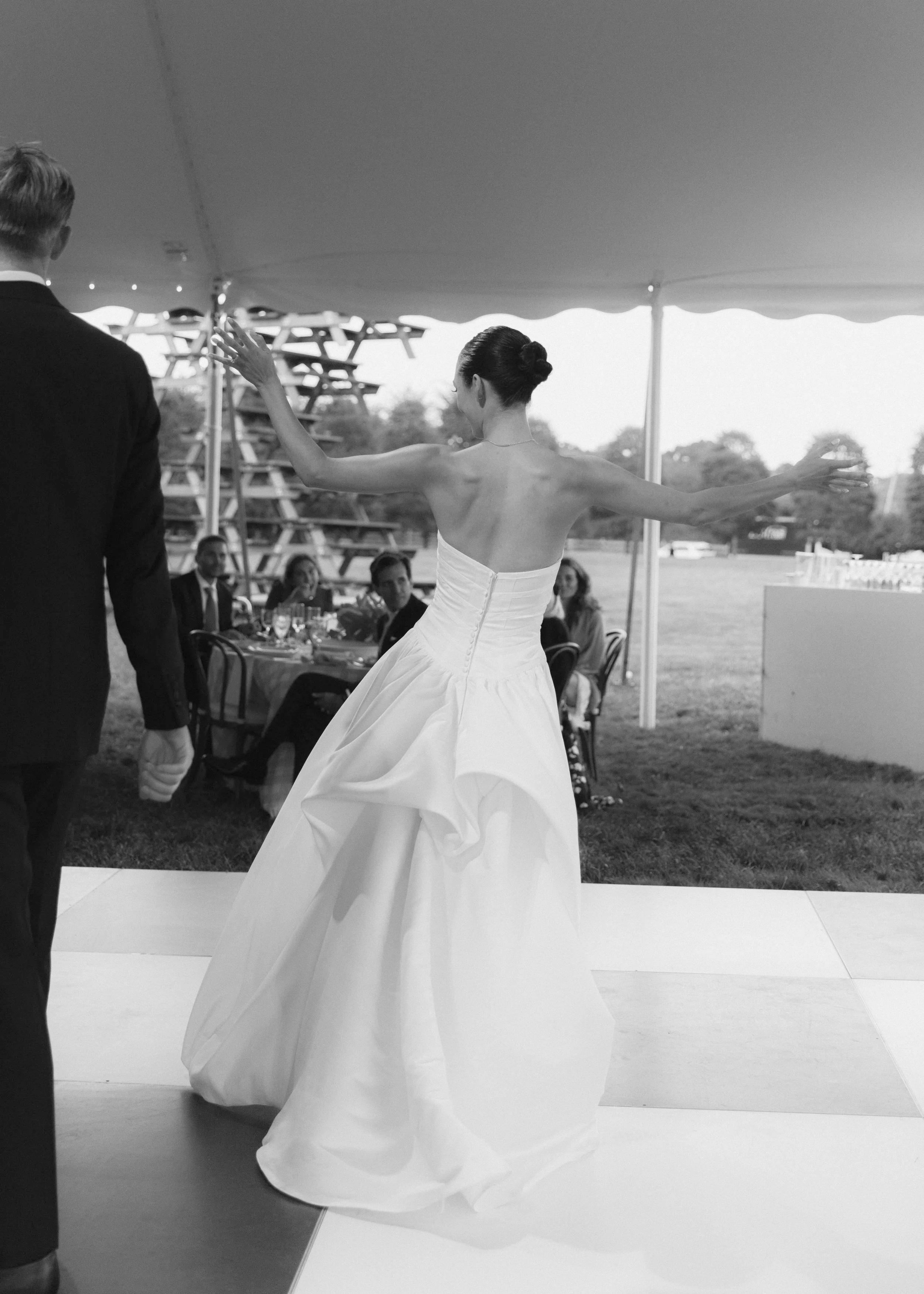 Black and white photo of a bride in a strapless gown dancing with arms outstretched on a checkered floor, with a man in a suit nearby and guests seated in the background.