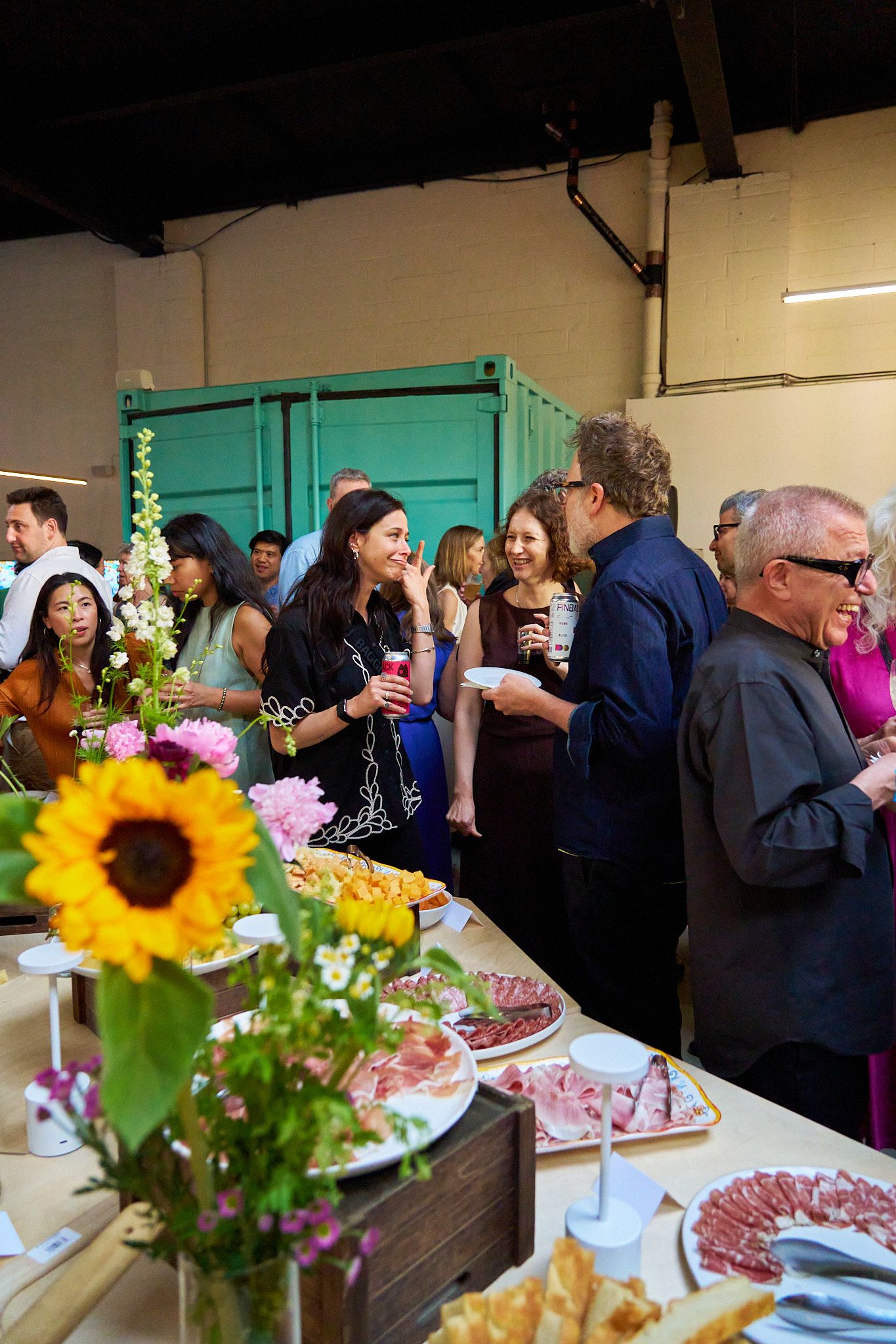 An indoor gathering with people mingling around a food-laden buffet table adorned with flowers, including a prominent sunflower.