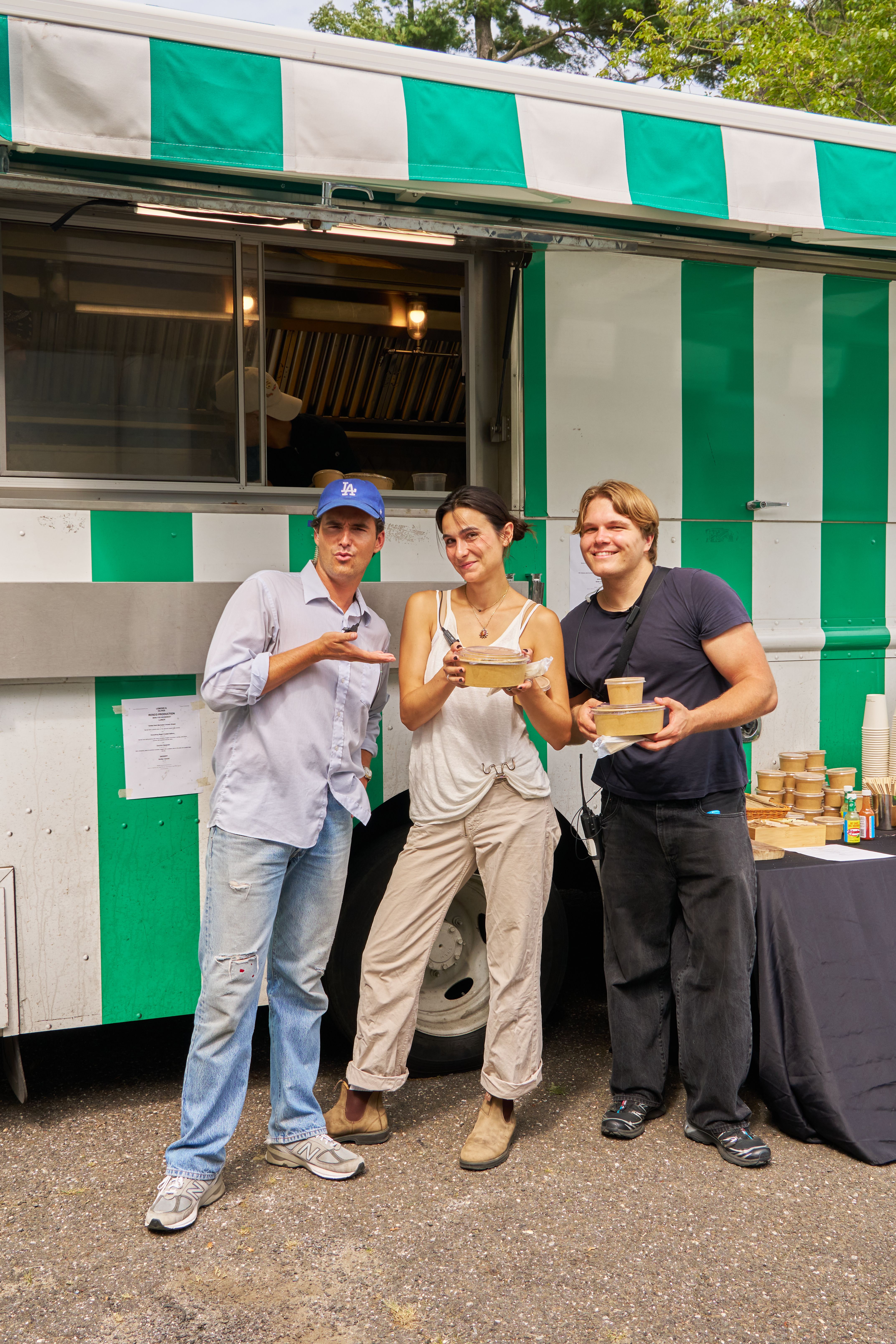 Three people, two men and a woman, smile and hold food containers in front of a green and white striped food truck.