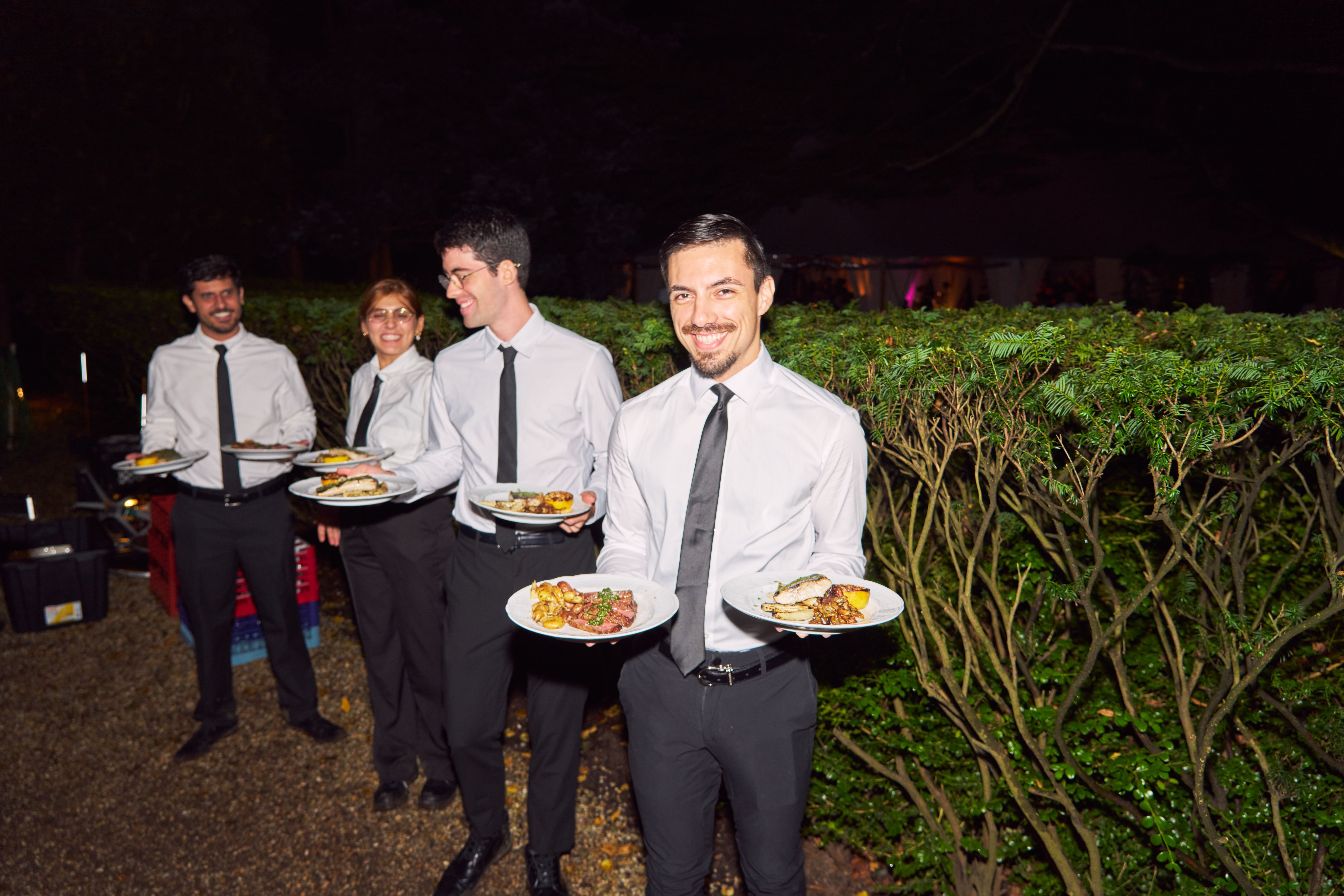 Four smiling servers in uniforms holding plates of food outdoors at night.