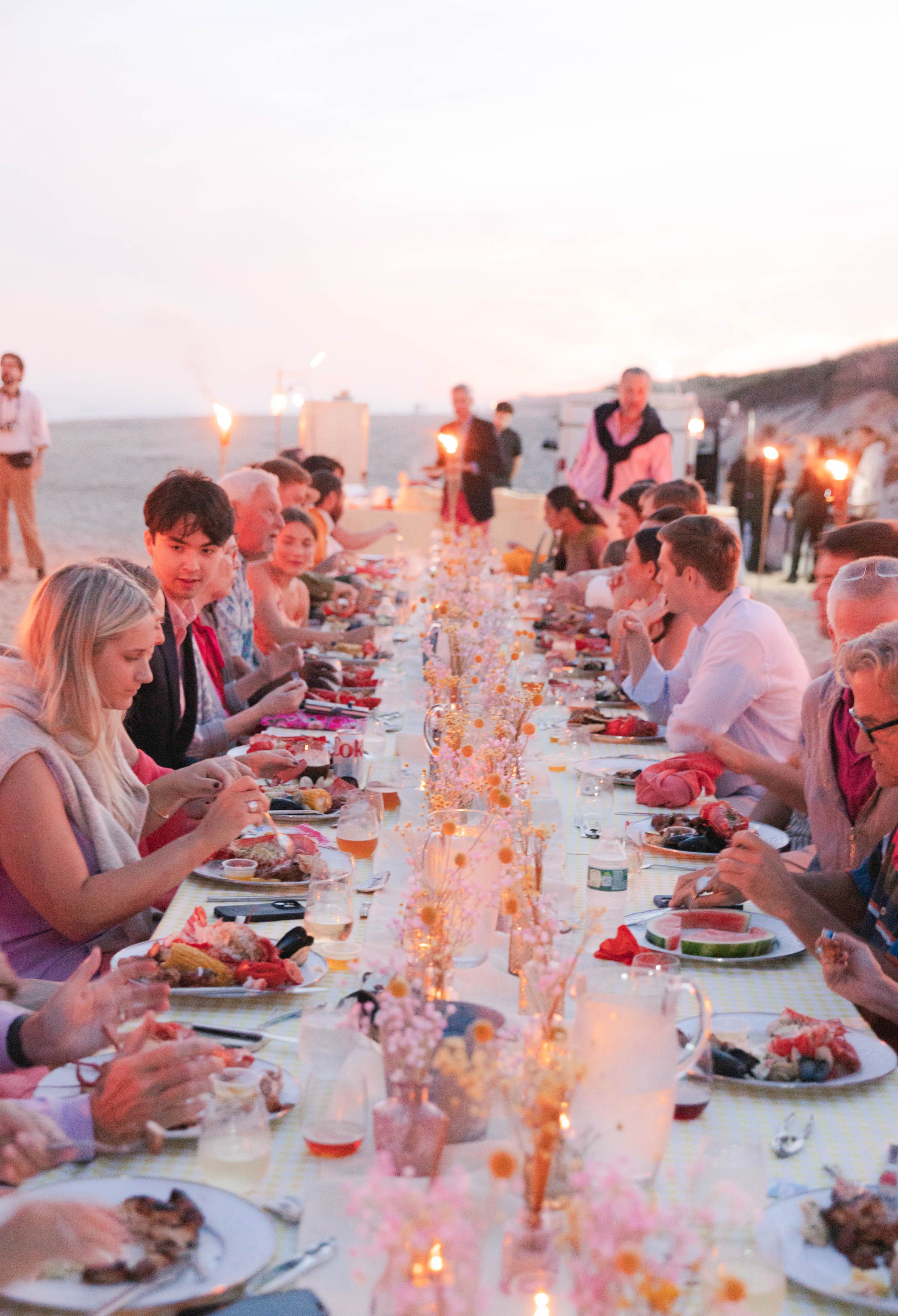 Guests dine at a long, decorated table on a beach during a pink-hued sunset.