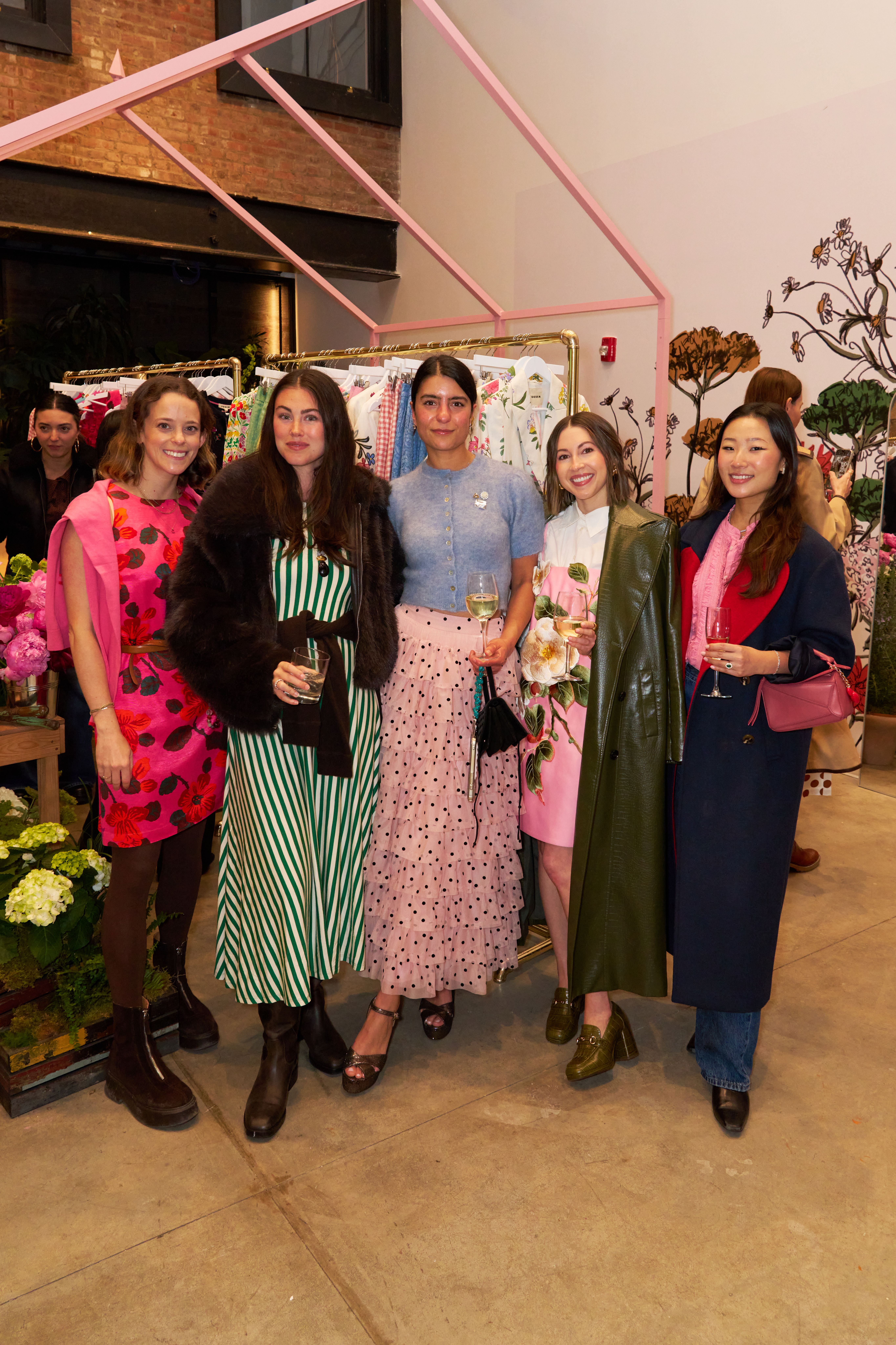 Five women in colorful, fashionable attire smile in a retail space with clothing racks and floral decor.
