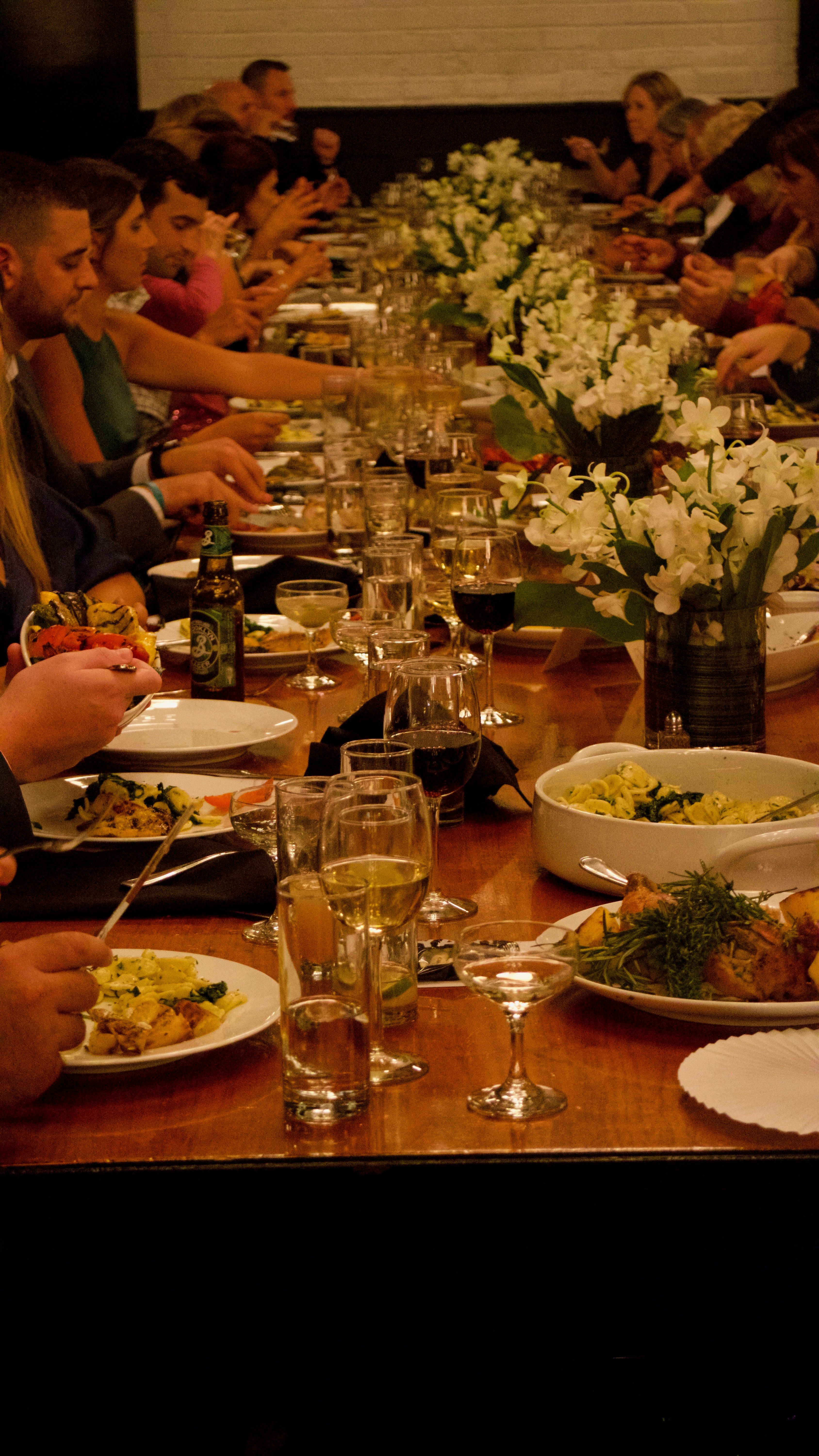Guests enjoying a long-table communal dining experience with plated dishes, wine, and floral arrangements.