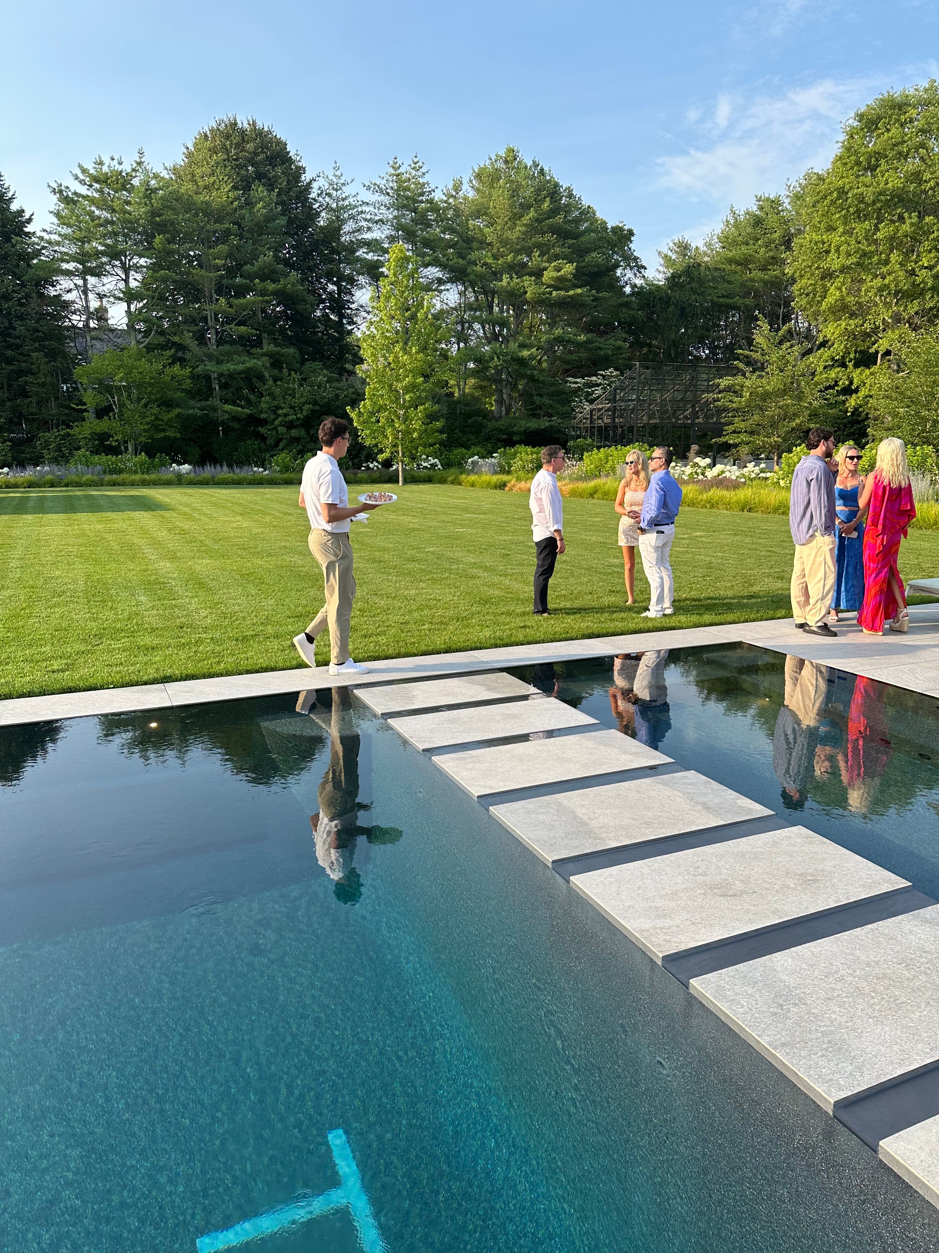 A male server walks on stepping stones over a reflective pool, holding a tray, in a modern garden.