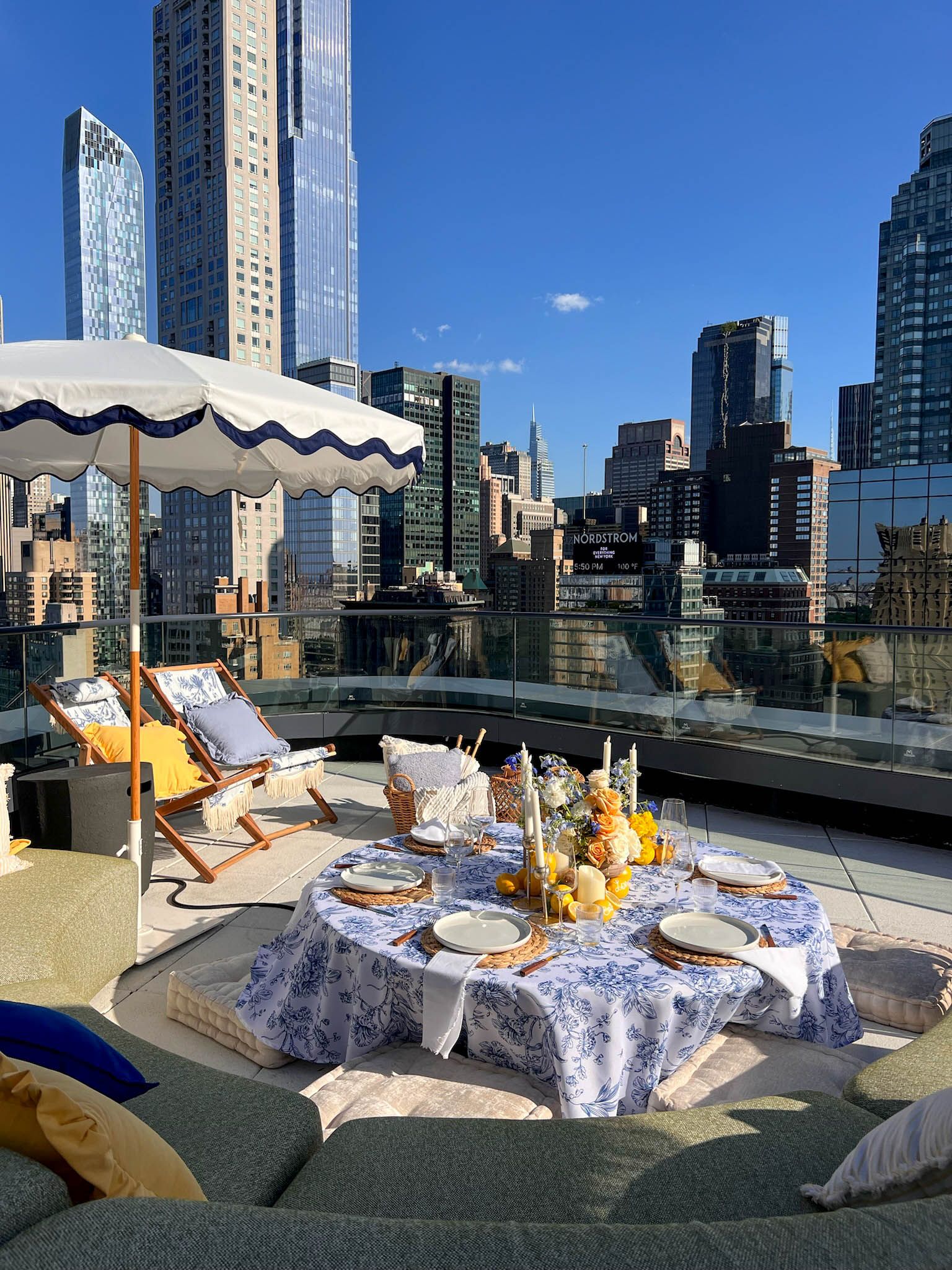An elegant rooftop picnic set with blue and white toile linens, a floral centerpiece, and a city skyline view.