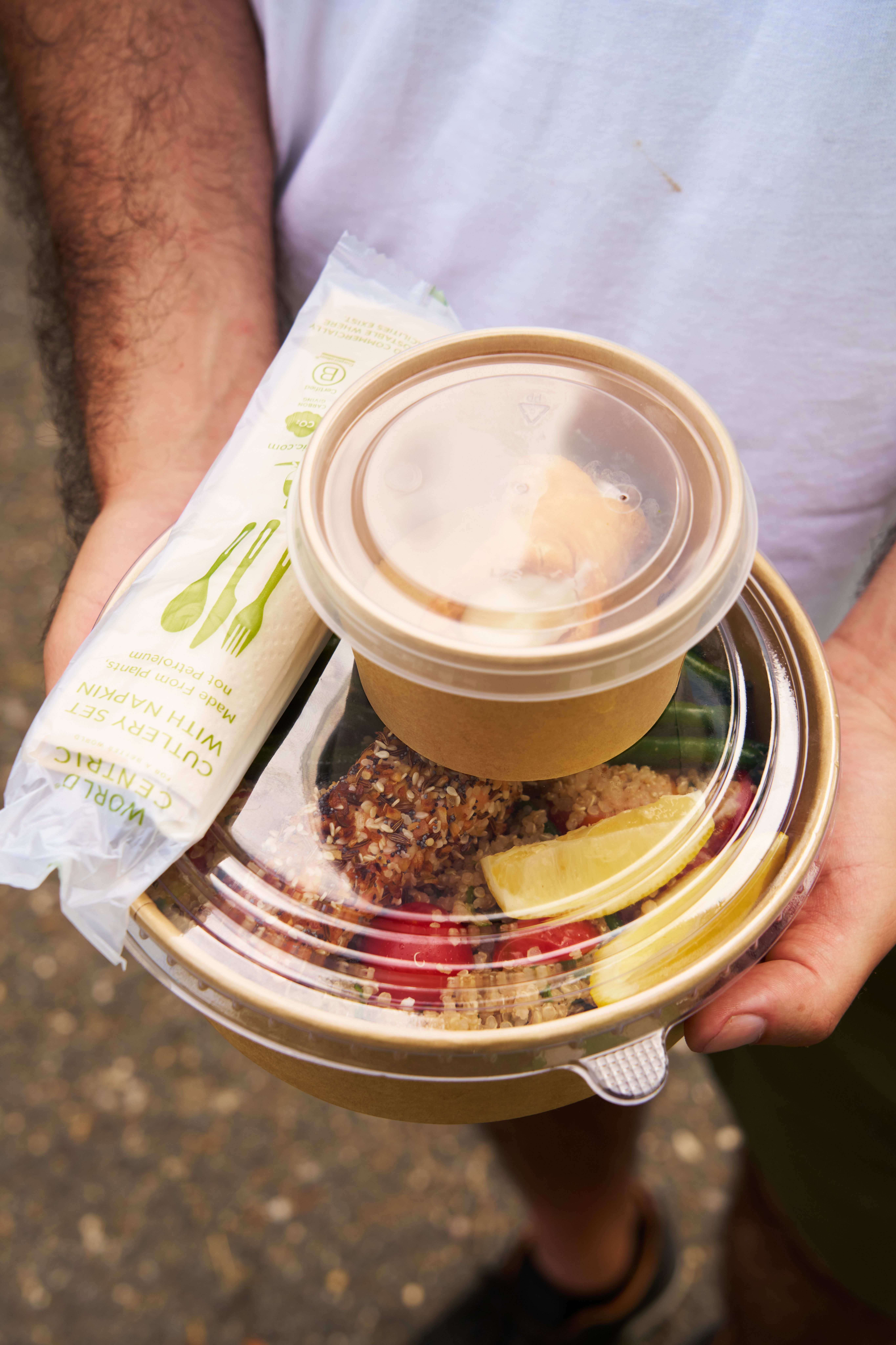 A person holds World Centric takeaway food in brown containers, featuring a main dish with fish, quinoa, and vegetables, a smaller side, and a plant-based cutlery set.