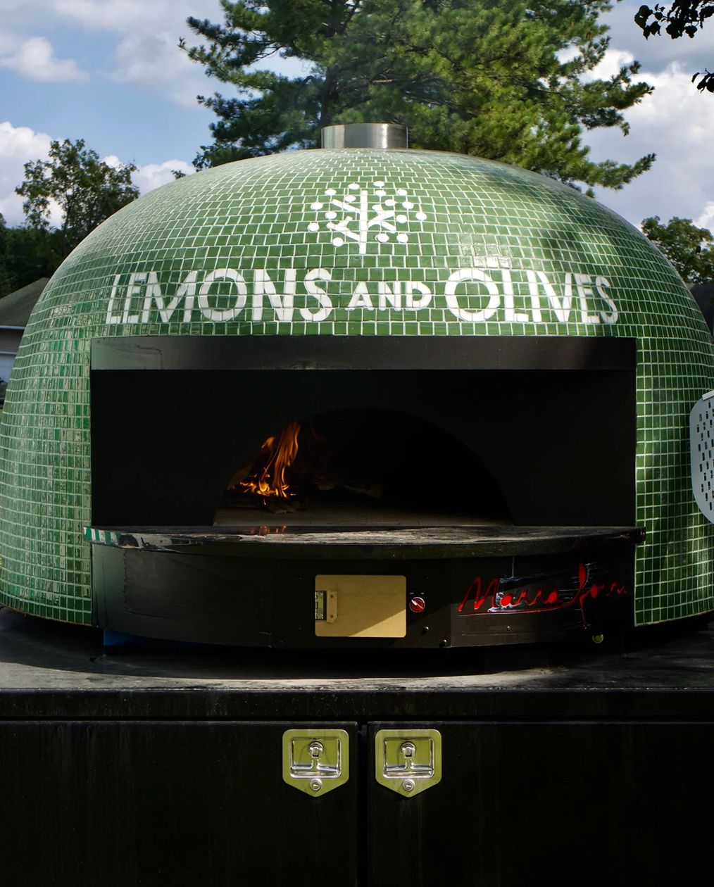 Green mosaic dome pizza oven with "Lemons and Olives" in white tiles and a fire burning inside.