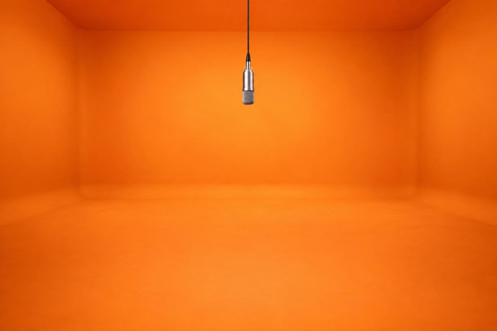 An empty tangerine cyclorama with one vintage condenser microphone hanging on a thin cable, locked camera, no furniture