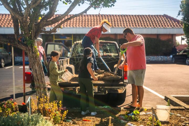 Students and adults filling buckets with garden material from the back of a pickup truck beside a planted median