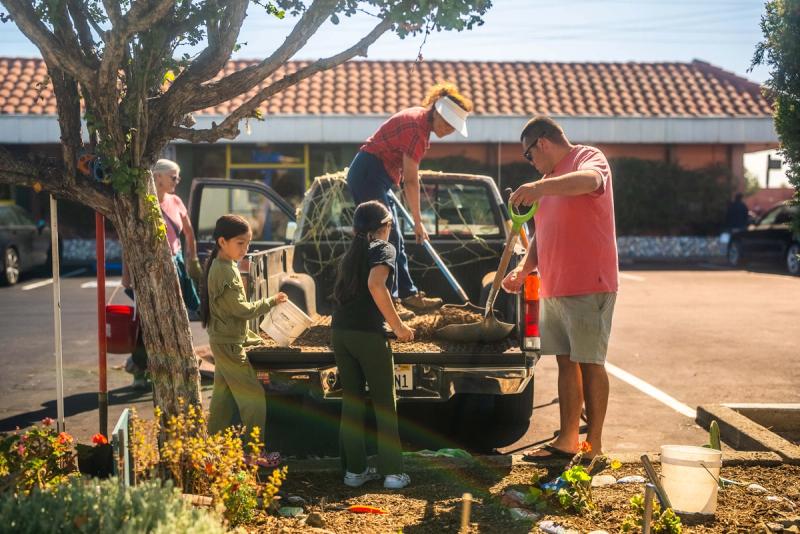 Students and adults filling buckets with garden material from the back of a pickup truck beside a planted median