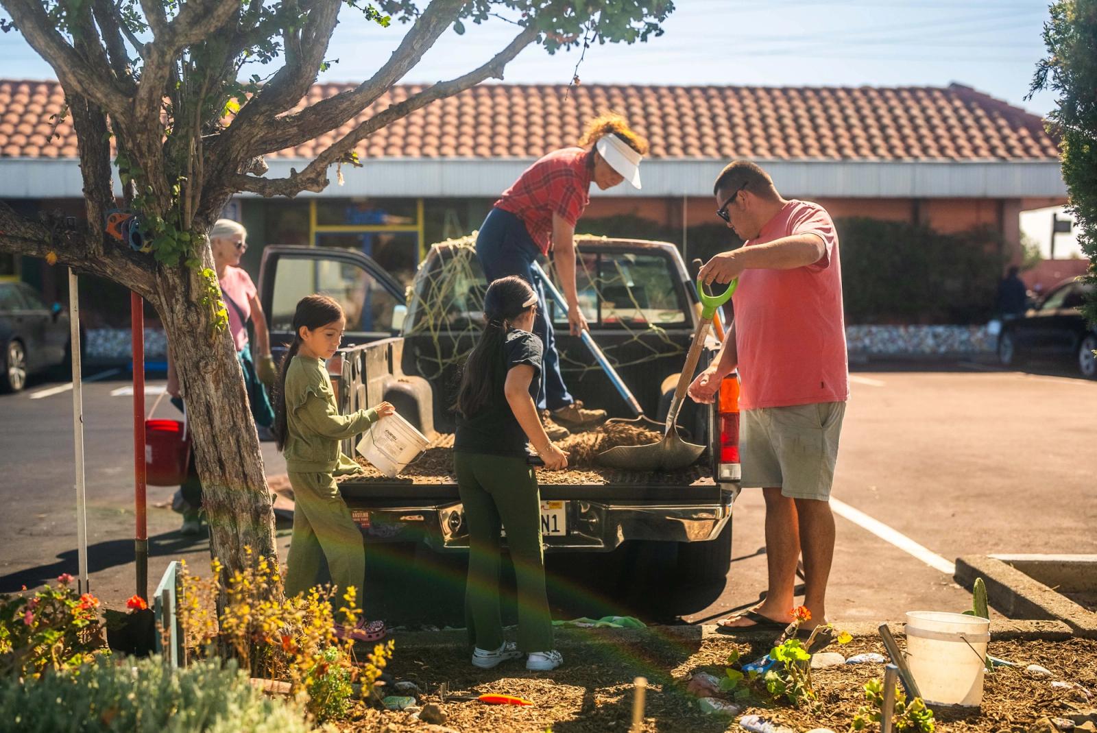 Students and adults filling buckets with garden material from the back of a pickup truck beside a planted median