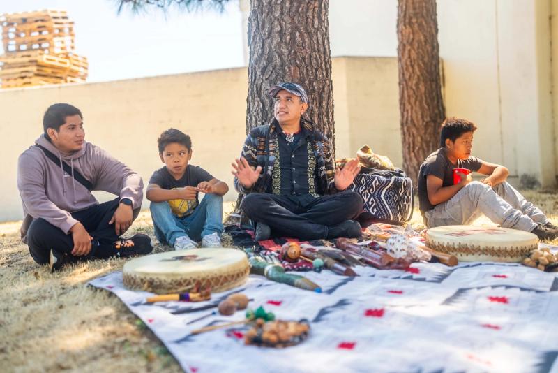 A gathering leader seated with students and an adult beside drums and instruments laid out on a patterned cloth