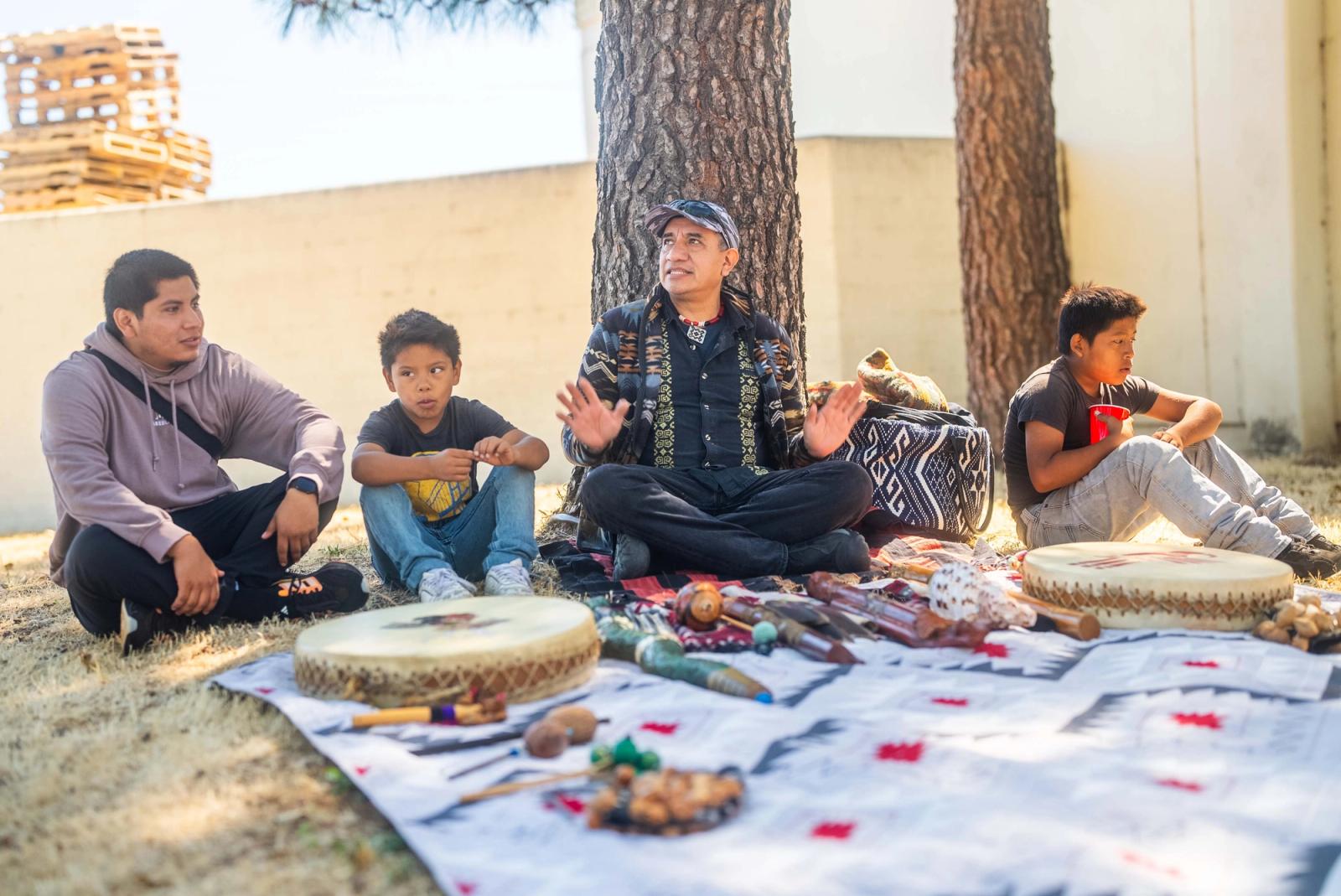 A gathering leader seated with students and an adult beside drums and instruments laid out on a patterned cloth