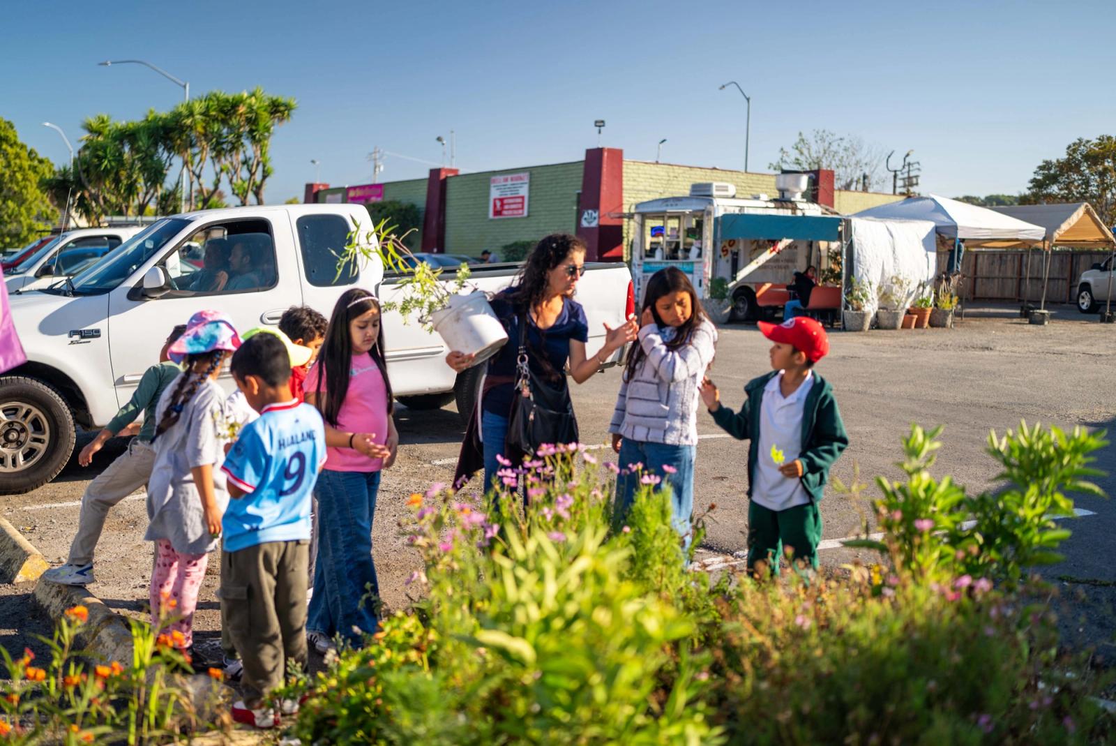 Students gathered with an adult beside a flowering garden along a neighborhood sidewalk