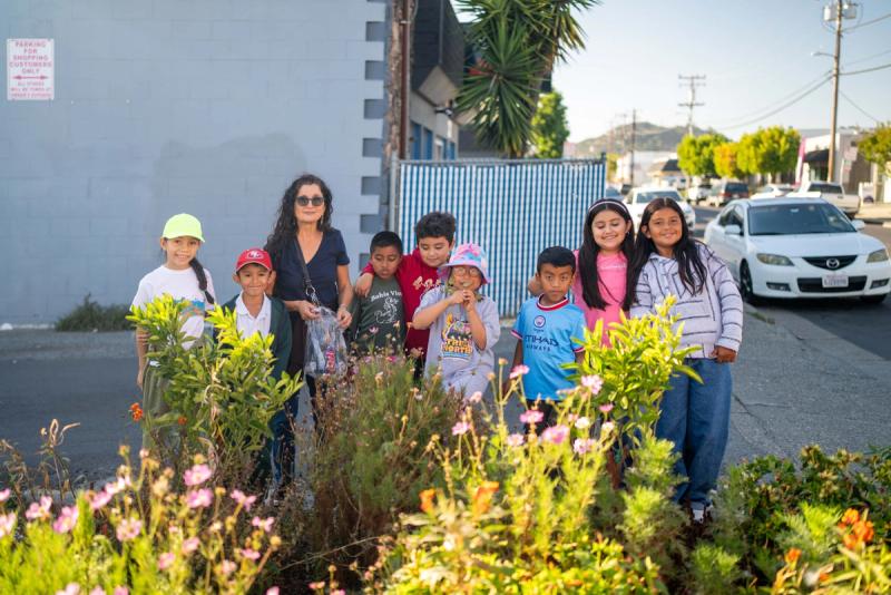 Young people and an adult standing among flowering plants in a neighborhood garden