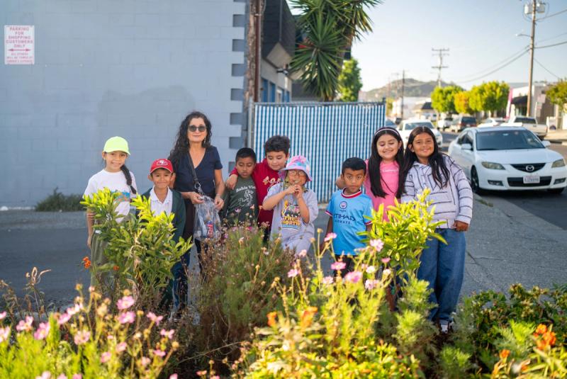 Young people and an adult standing among flowering plants in a neighborhood garden