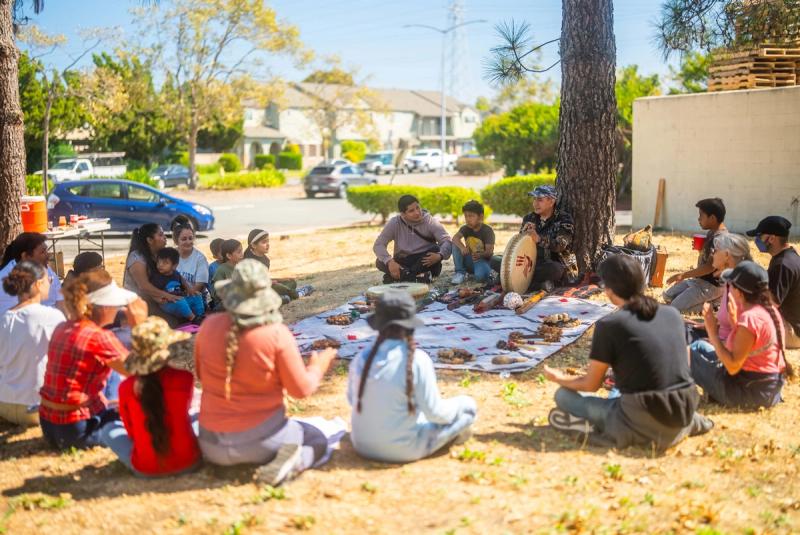 Students and adults gathered in a circle outdoors beneath the trees