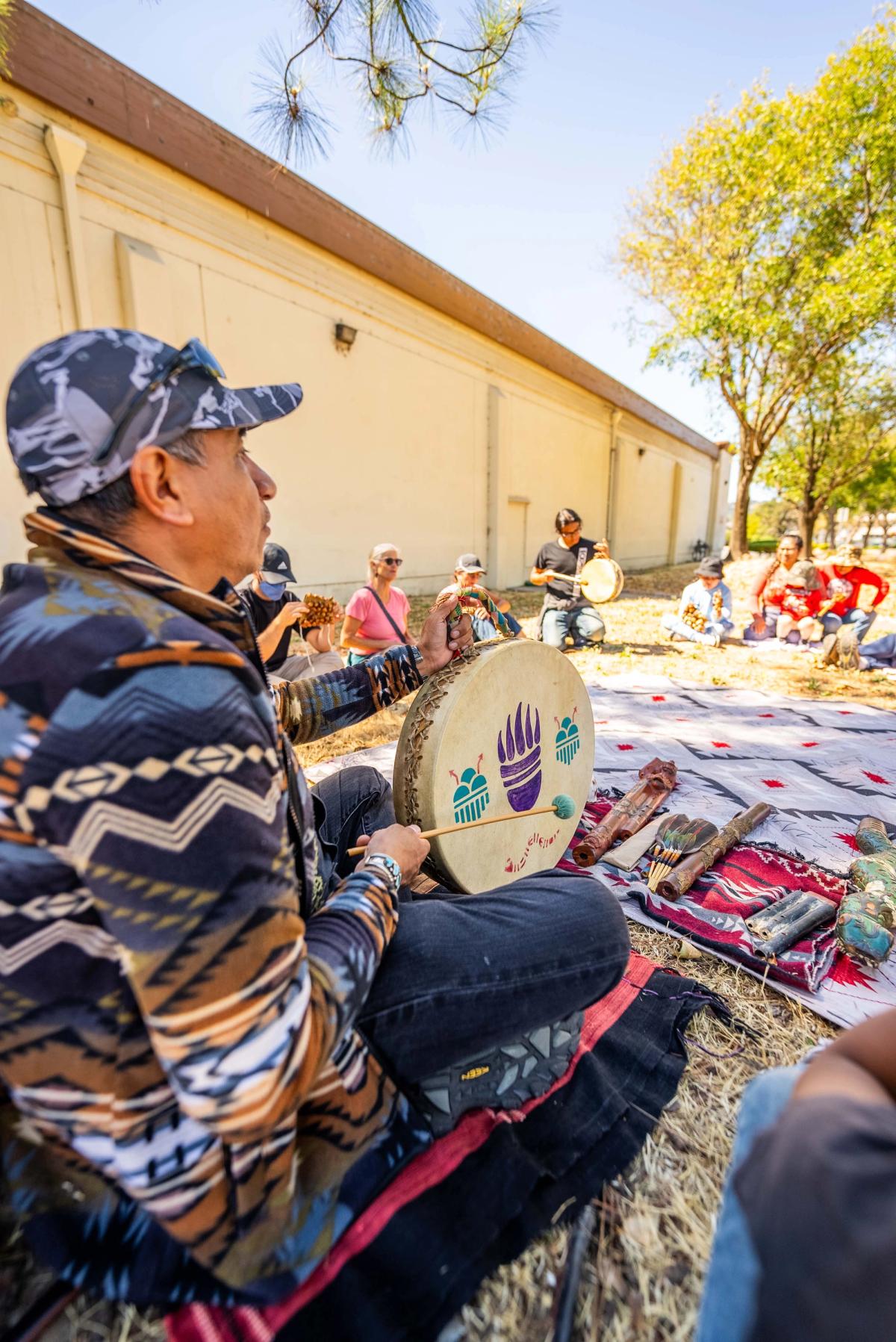 A gathering leader playing a hand-held drum with other instruments laid out beside him and participants seated nearby