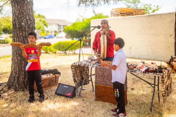 An adult and a young person standing before an outdoor gathering beside drums