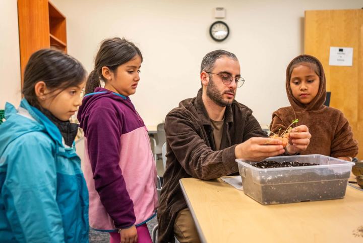 An adult showing a seedling to students gathered around a container of soil