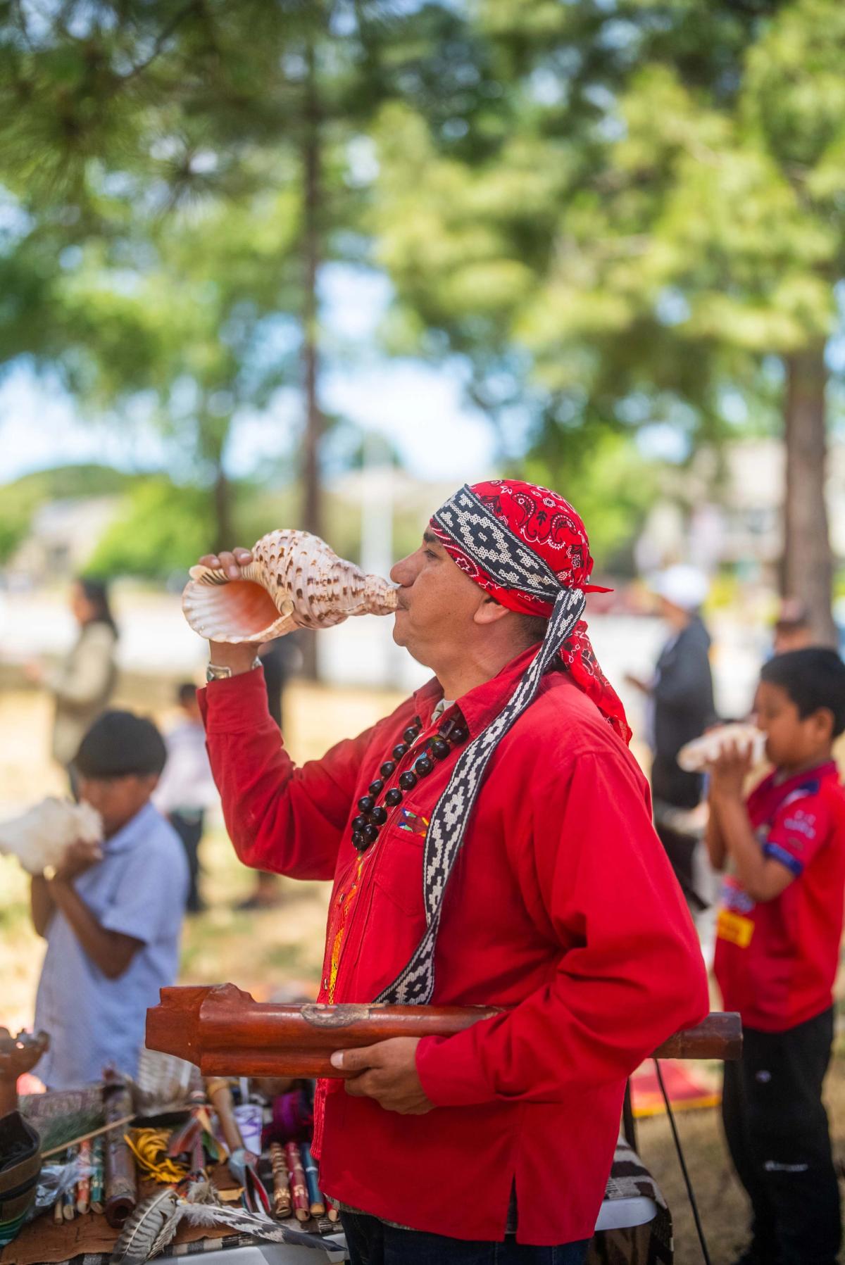 A gathering leader in a red shirt playing a large shell outdoors with young people nearby