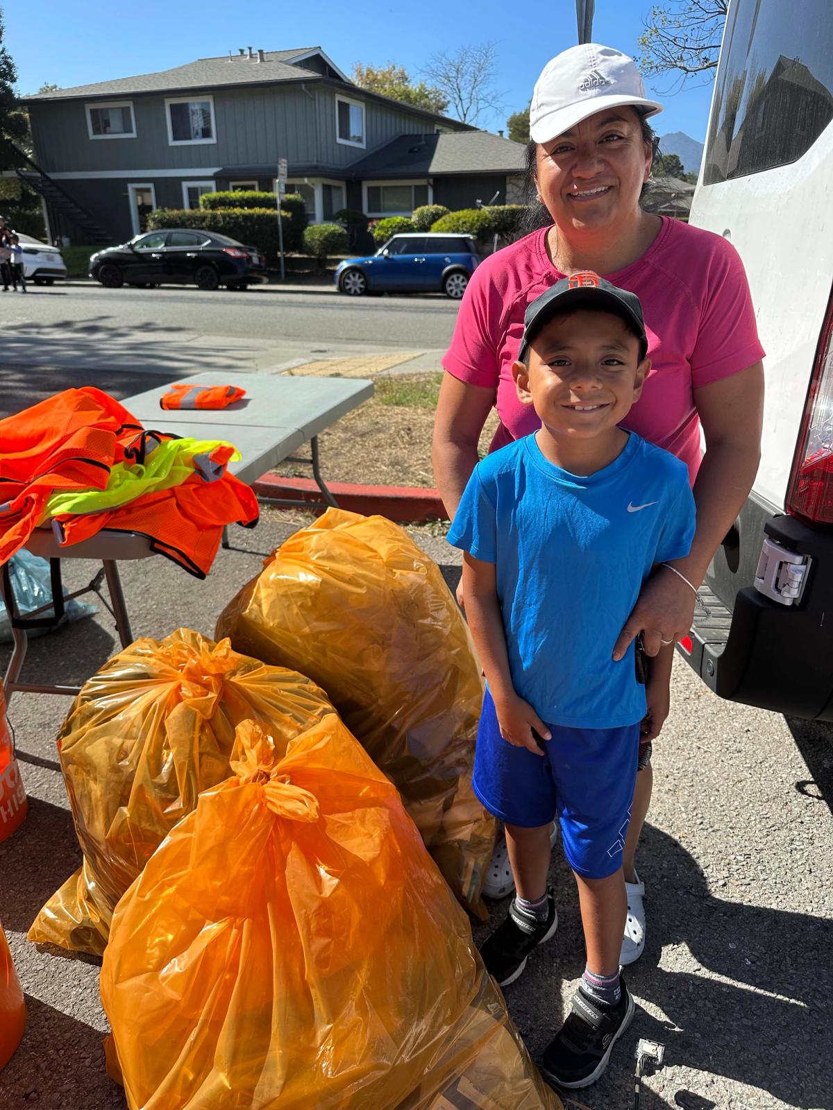 A child and an adult smiling beside collected trash bags, with reflective vests on a nearby table