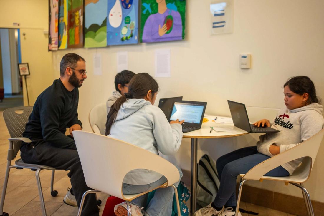 An adult supporting students working with notebooks and laptops at a classroom table
