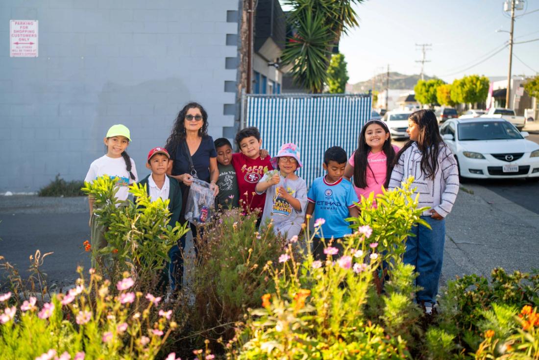 Students and an adult standing behind flowering plants in a neighborhood garden