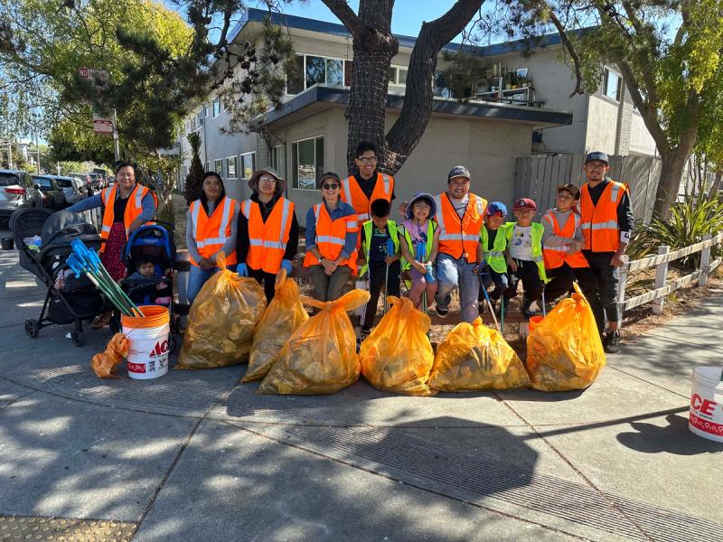 Children and adults in reflective vests standing behind bags of collected trash on a neighborhood sidewalk
