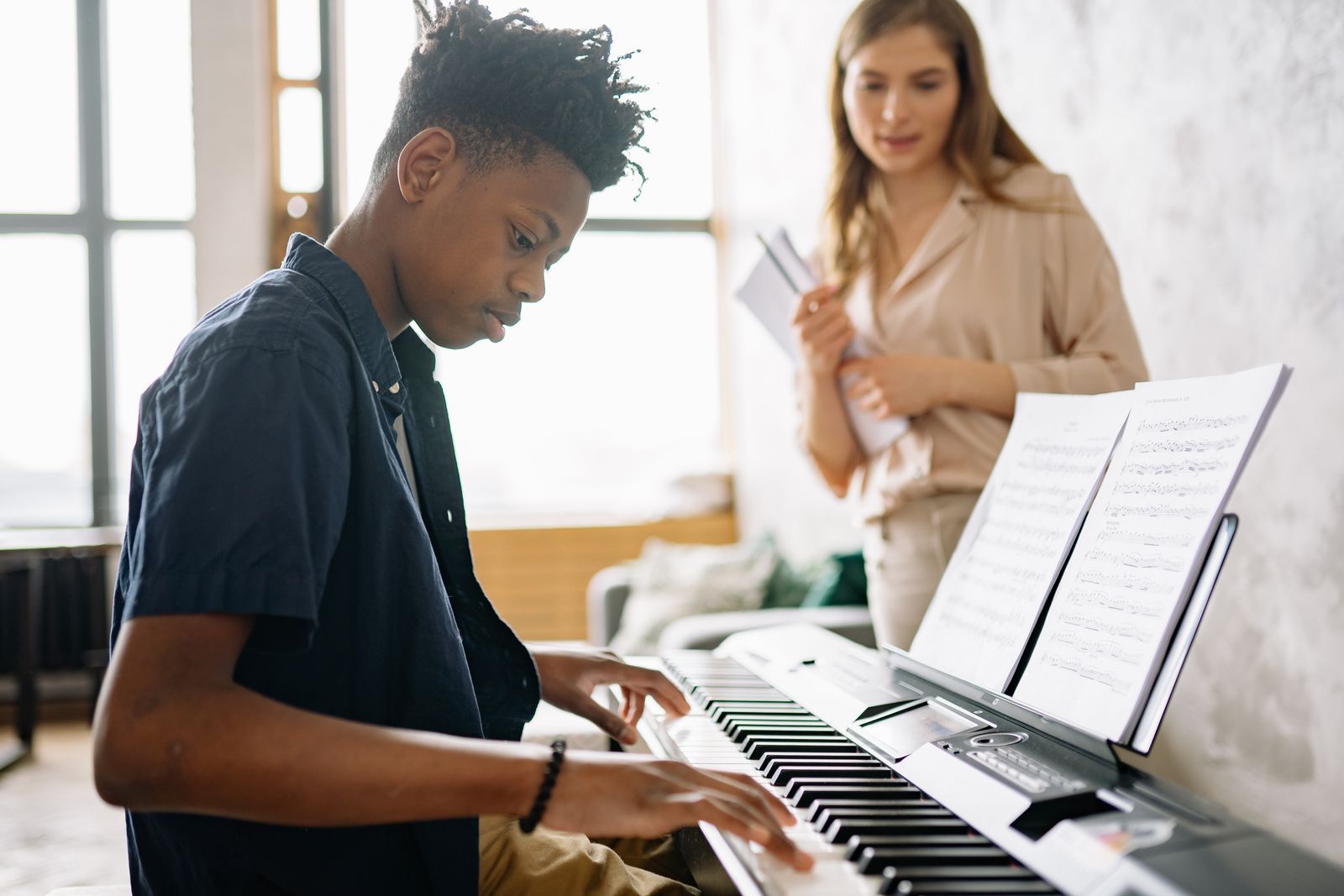 Adolescent jouant du piano numérique avec une partition, professeure en arrière-plan
