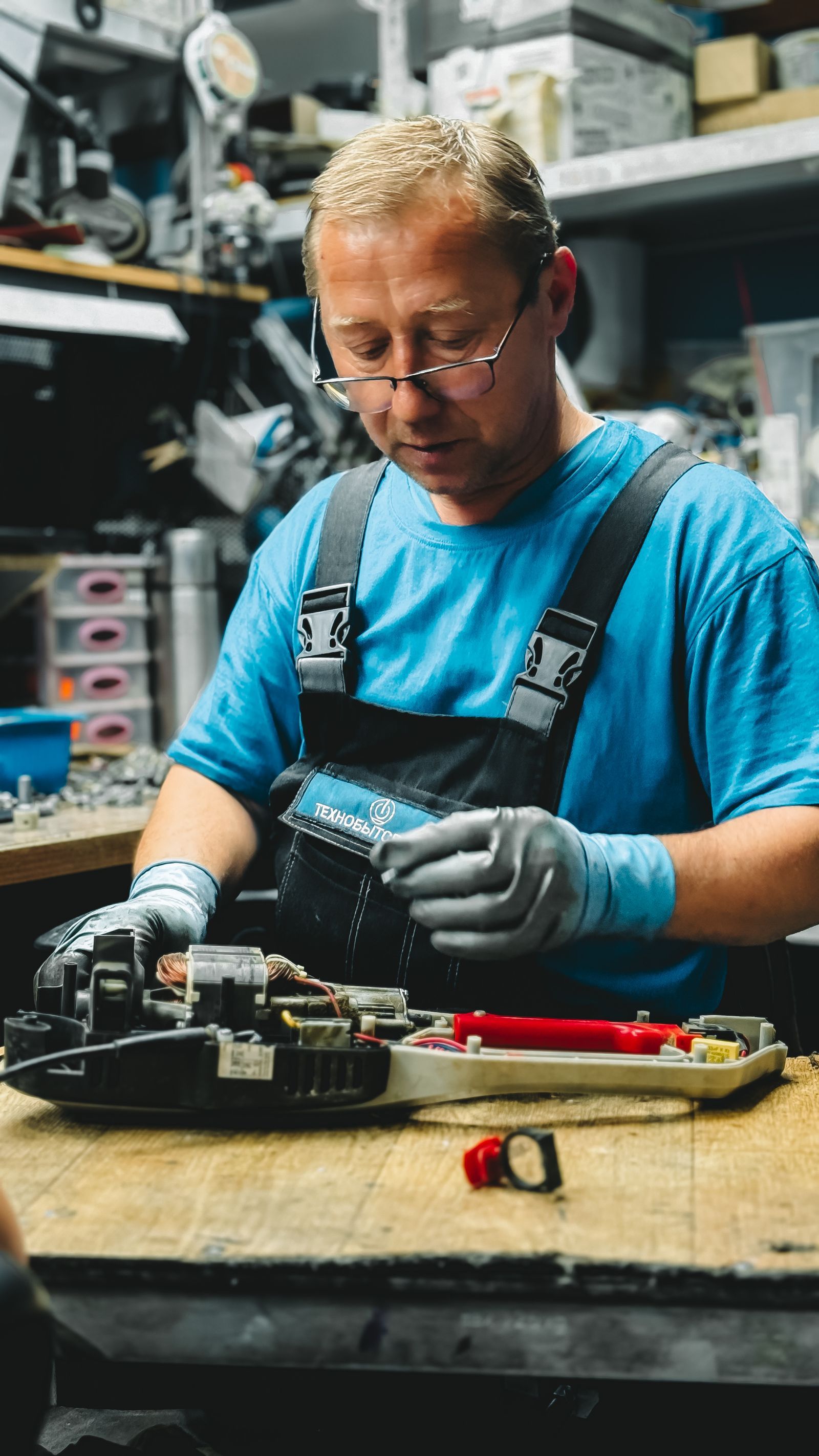 Technicien en tenue de travail bleue assis à un établi, équipé de gants et d'outils de précision