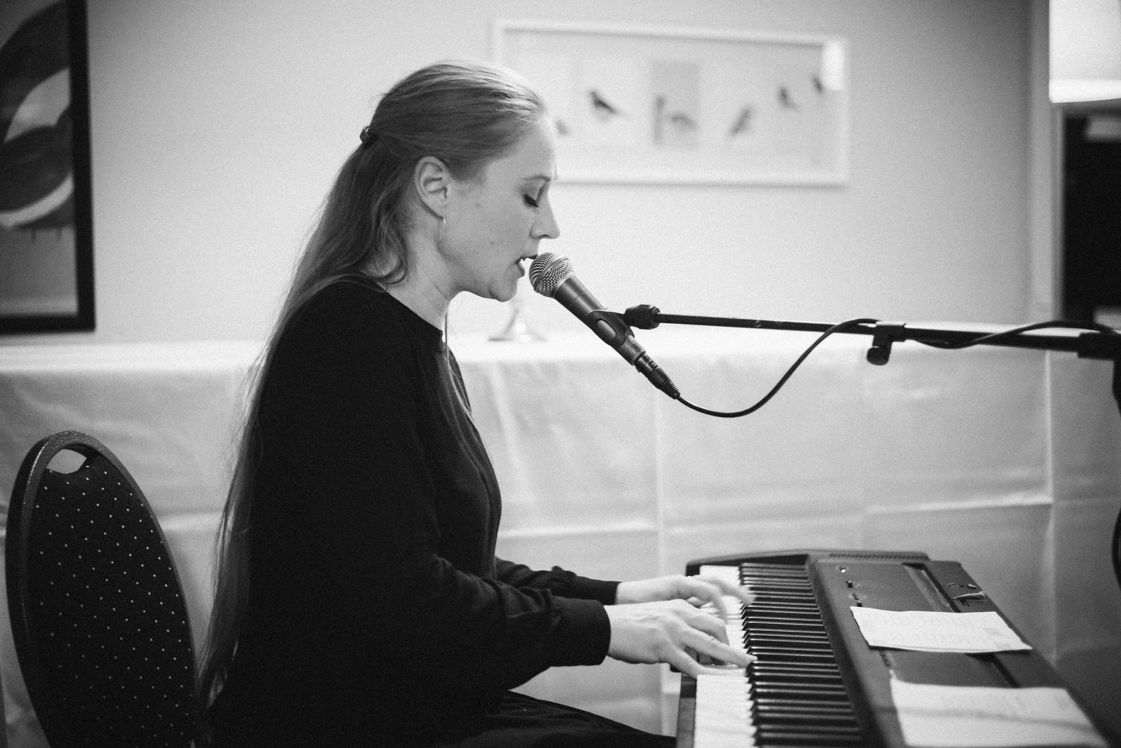 Chanteuse au micro, penchée sur un clavier électronique, dans une pièce claire, en noir et blanc