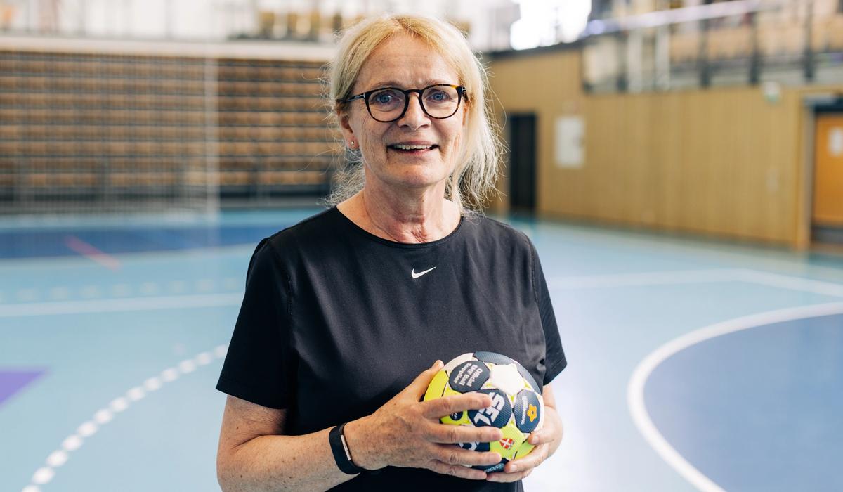 Dame med sort t-skjorte, lyst hår og briller som står med en håndball i hendene og smiler inn i kameraet 