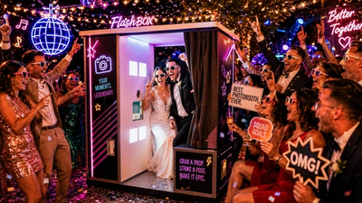 Ein lächelndes Hochzeitspaar in einer Fotobox umgeben von feiernden Gästen.
