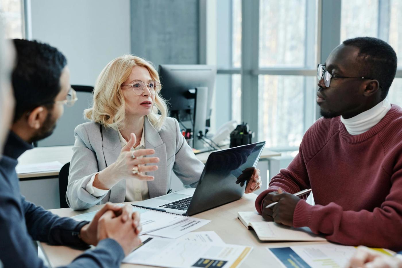 Three colleagues discuss documents at a table with a laptop.