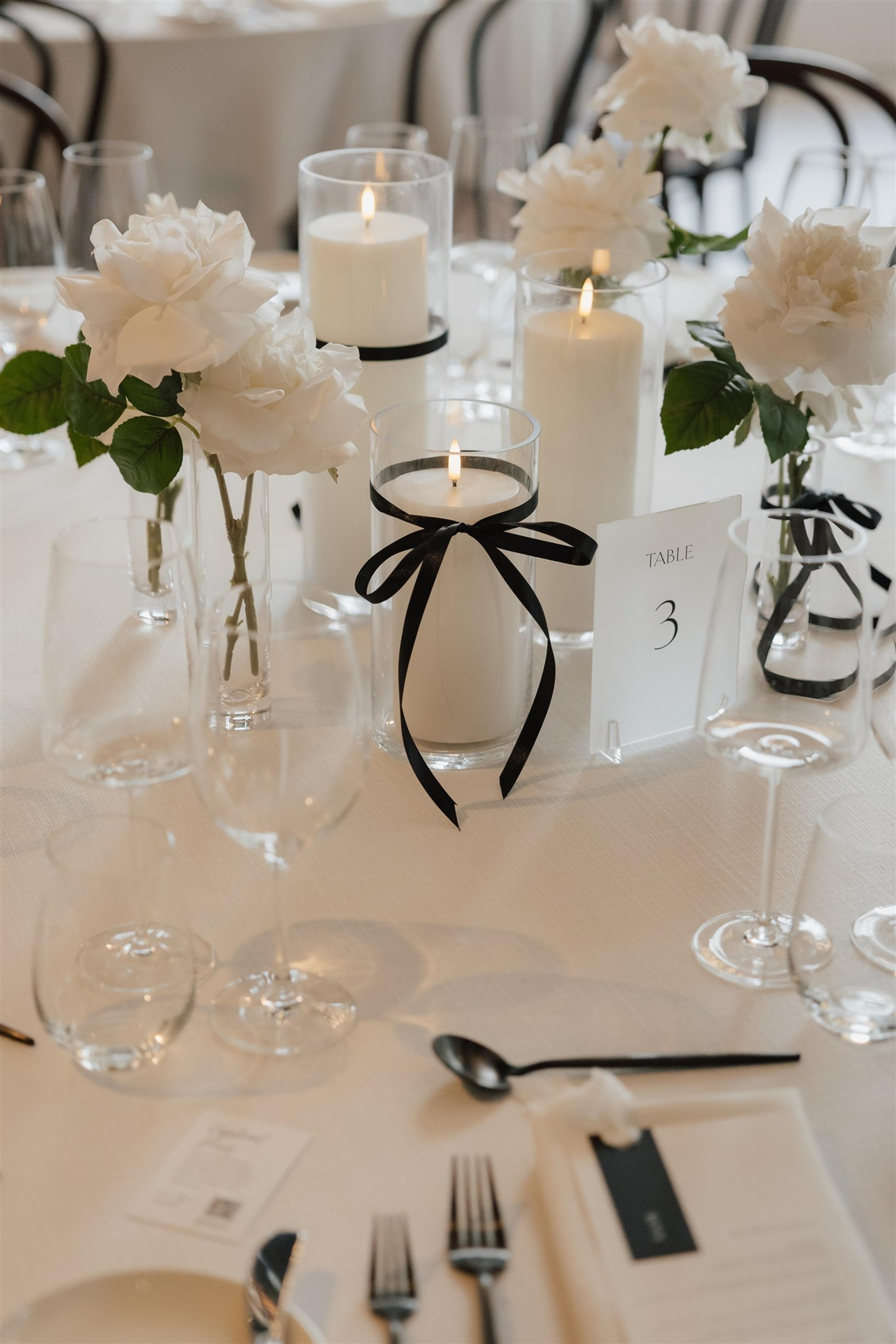 Close-up of table three styling with white roses, glass candle holders and black ribbon details at The Valley Estate Gold Coast wedding.